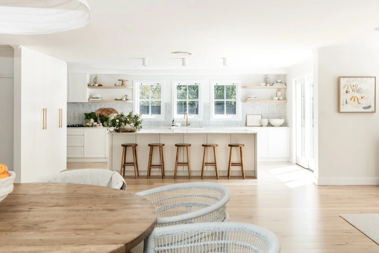 Bright, white kitchen with three windows, wooden bar stools at a white kitchen island, open shelves with decor, and a round wooden dining table with wicker chairs.