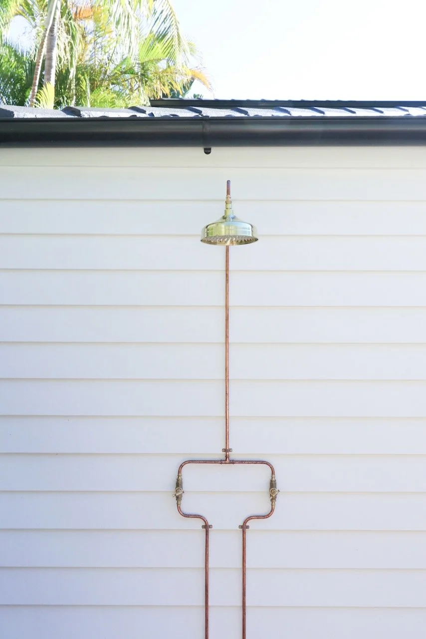 An outdoor shower with a brass showerhead mounted on a white wall, with exposed copper pipes connecting to it.