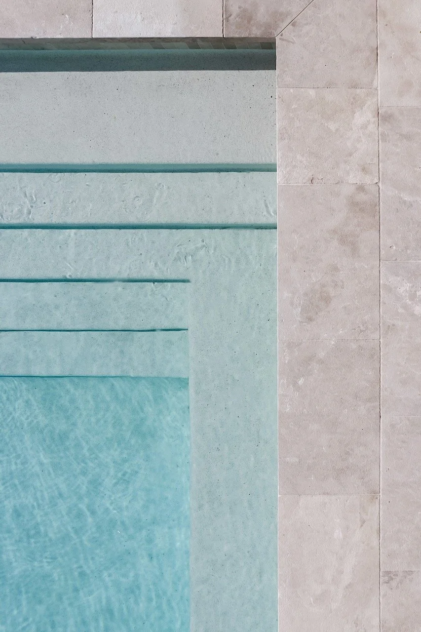 Close-up of a swimming pool with light blue water, with steps leading into the pool, and beige tiled floor on the side.
