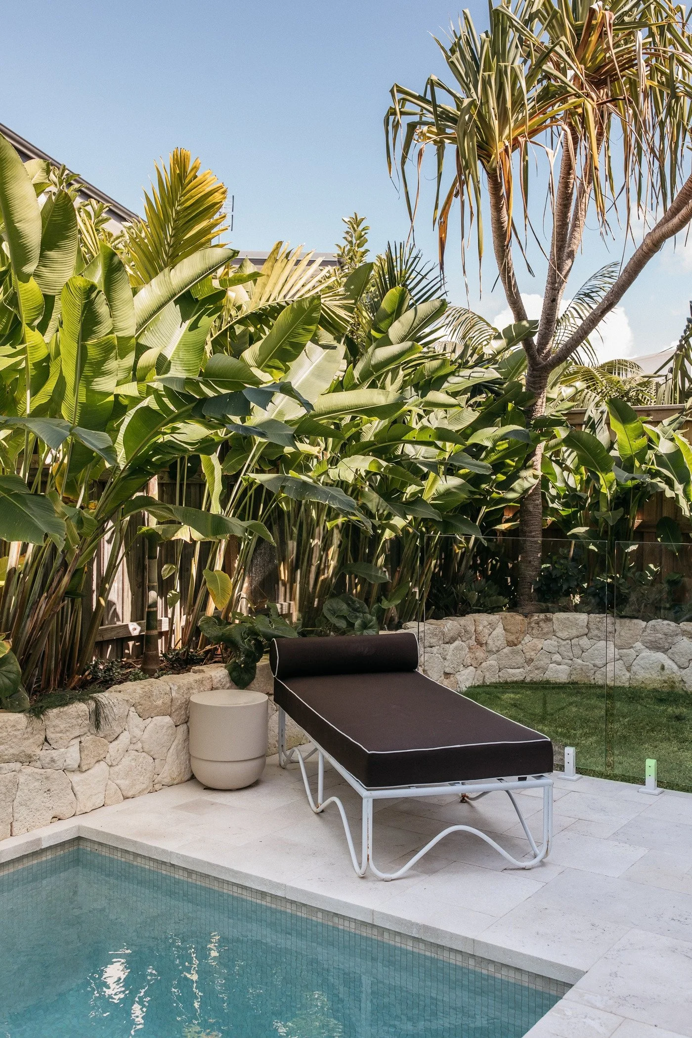 View of a backyard with a swimming pool, black lounge chair with white frame, white round side table, stone wall, lush green tropical plants, tall trees, and a clear sky.