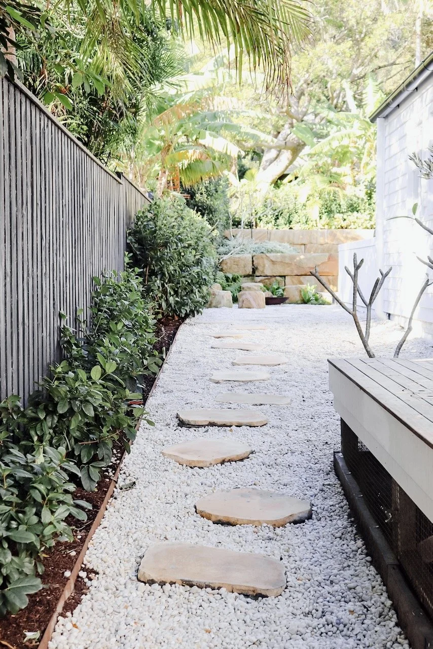 A gravel pathway with stepping stones, bordered by green plants and shrubs, leads to a garden area with rocks and trees in the background.