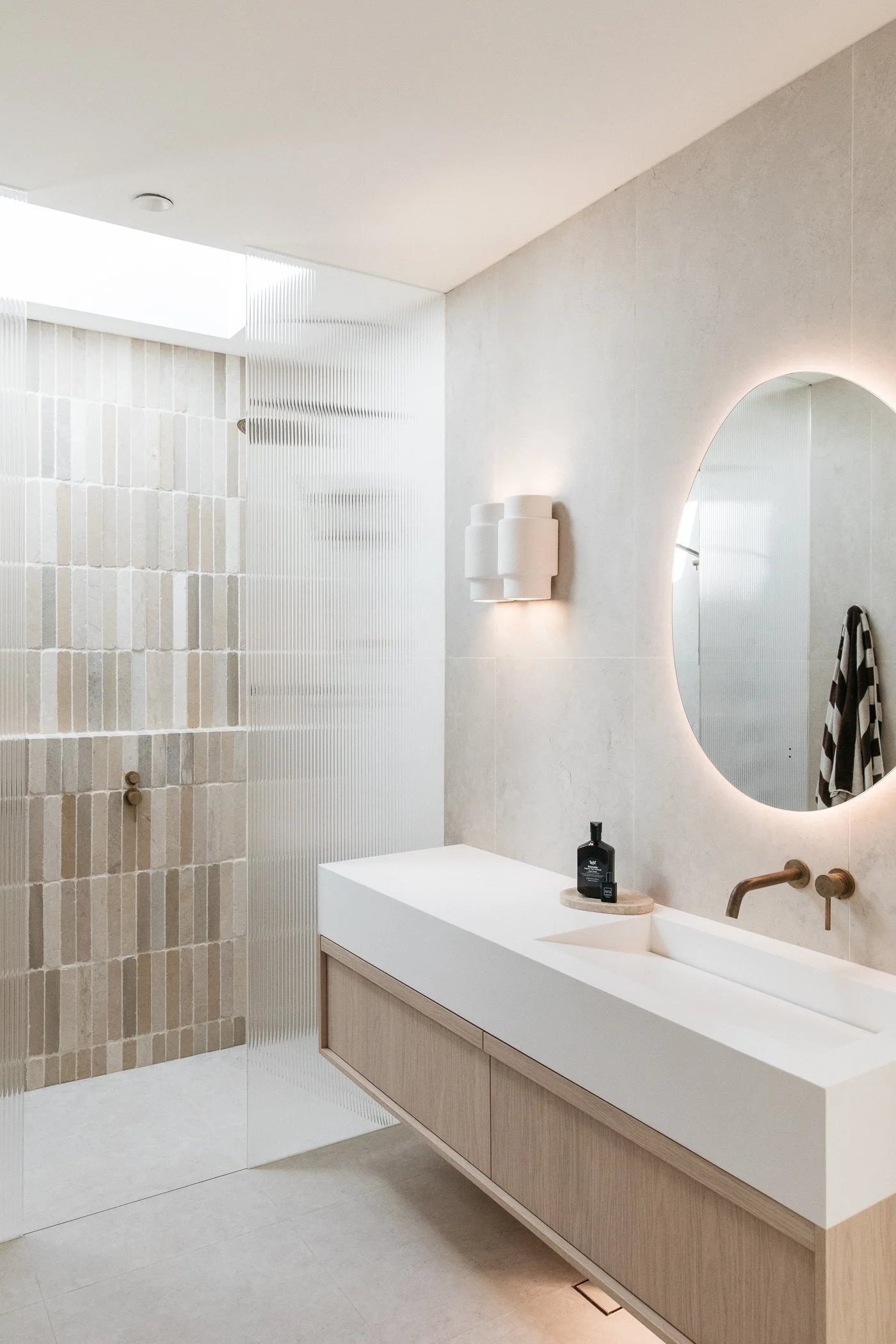 Modern bathroom with beige tiled walls and floor, a white vanity with wooden drawers, an oval mirror with backlighting, a white sink with a gold faucet, and a soap dispenser on the counter.