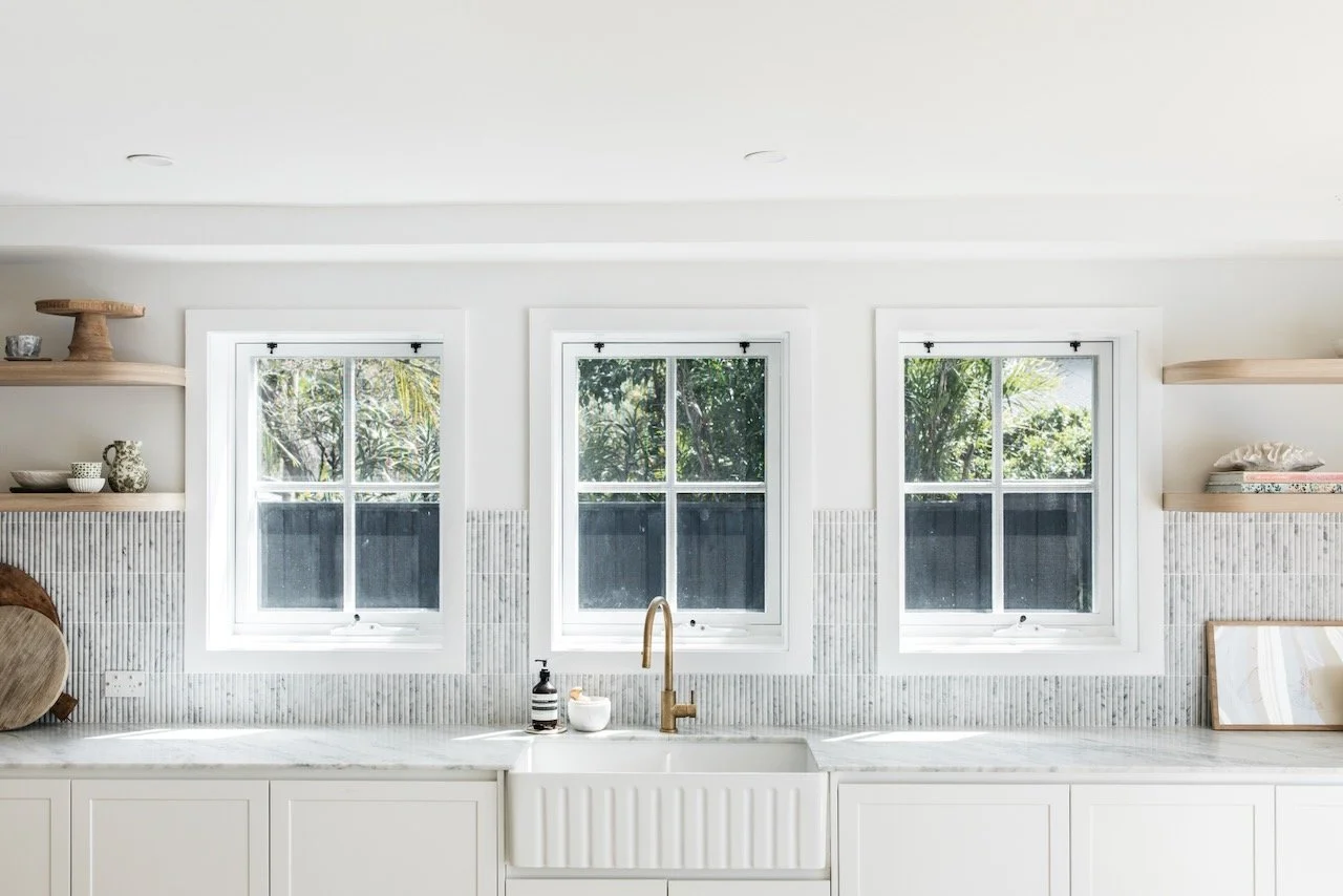 Bright, white kitchen with three windows behind a marble countertop and a white sink with a gold faucet. Decor includes open shelves with bowls, cups, a plant, and framed art.