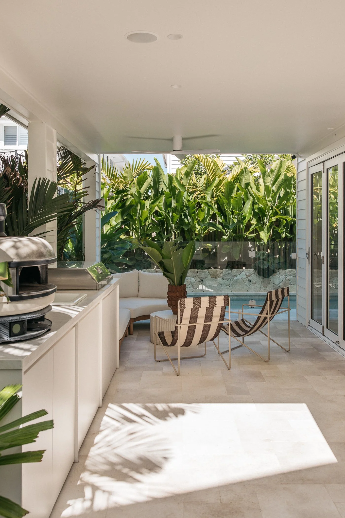 A white covered patio with a ceiling fan, lush green banana plants, a white outdoor sofa, striped patio chairs, a stone wall, and a swimming pool in the backyard.