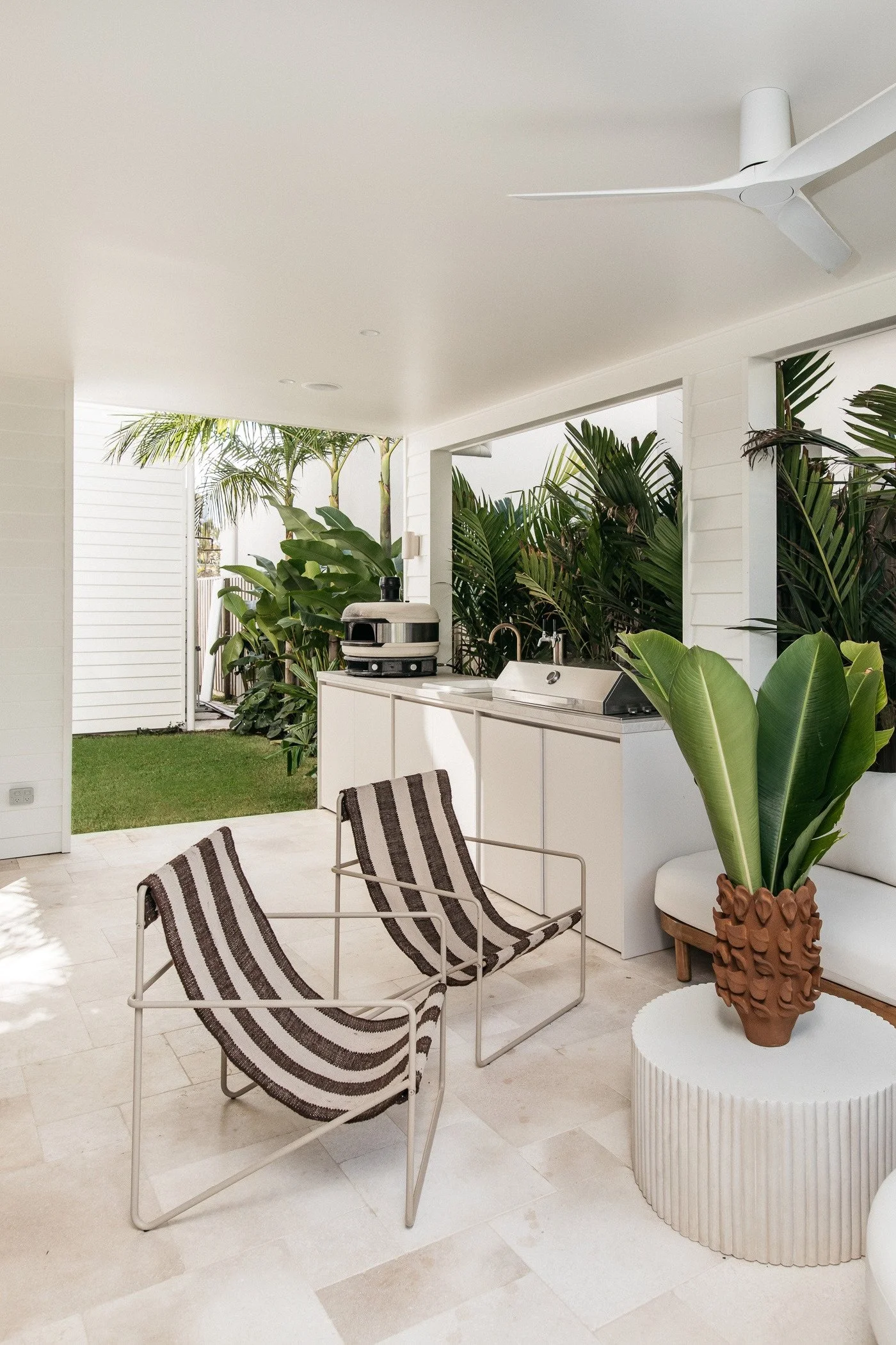 A modern outdoor patio with white walls and ceiling, tropical plants, a potted plant with large green leaves on a white ribbed side table, a stainless steel barbecue grill, a white counter, and chairs with black and white striped fabric. A ceiling fa