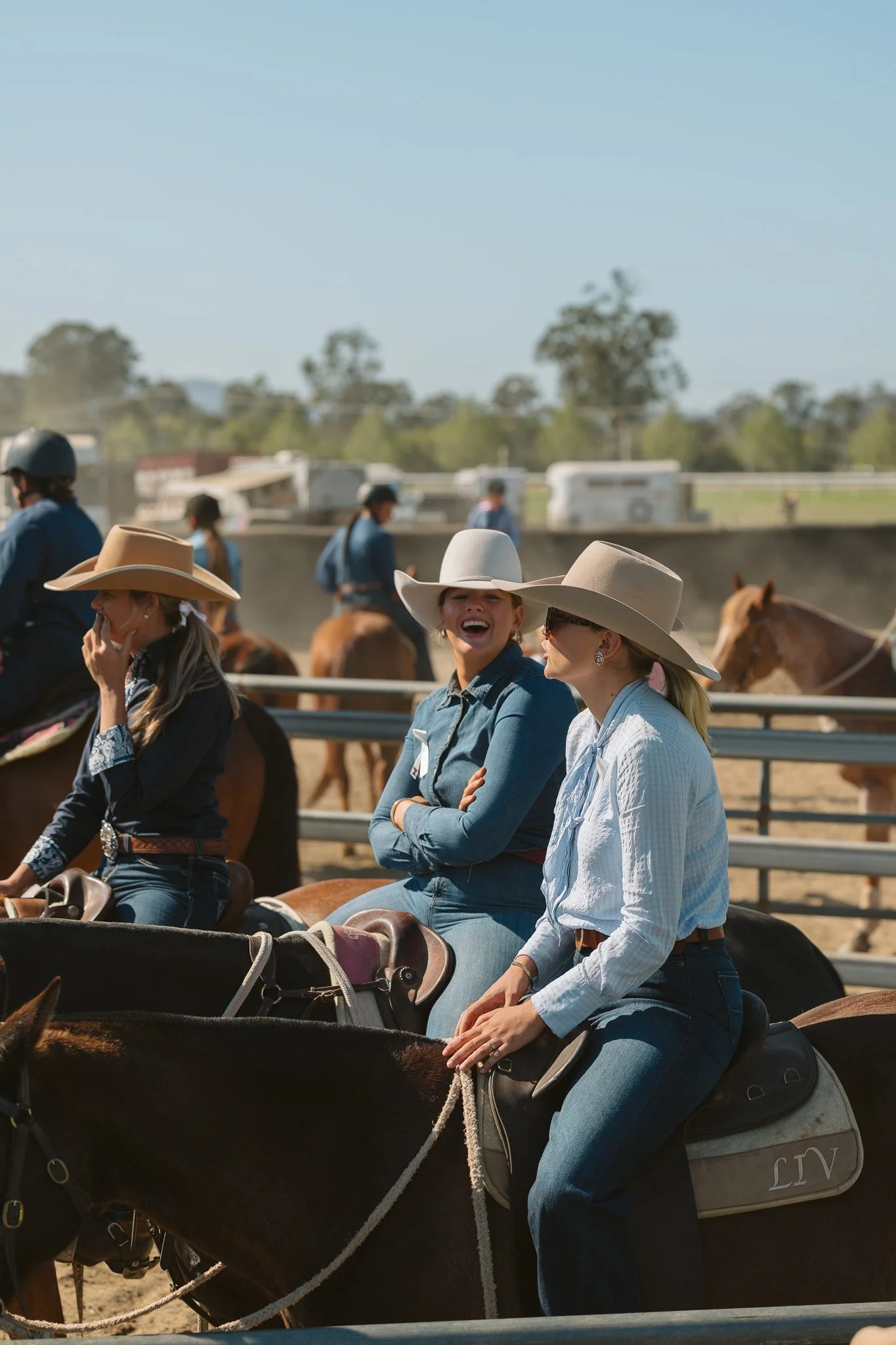 People riding horses at a rodeo, wearing cowboy hats and jeans, with a dirt arena and trailers in the background on a sunny day.