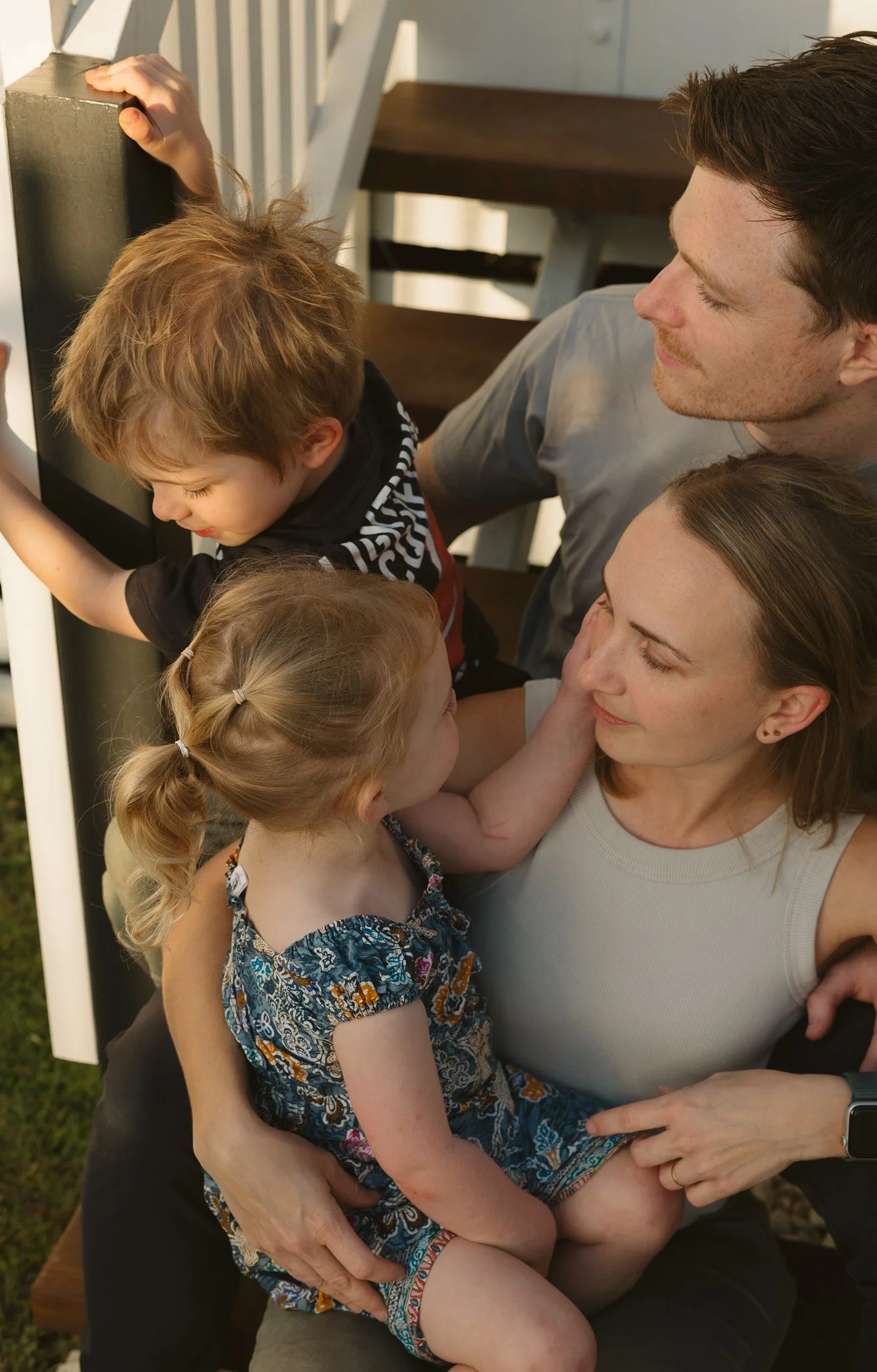 Family of five embracing outdoors near a staircase, including a woman, a man, two young girls, and a boy, with sunlight and greenery around.