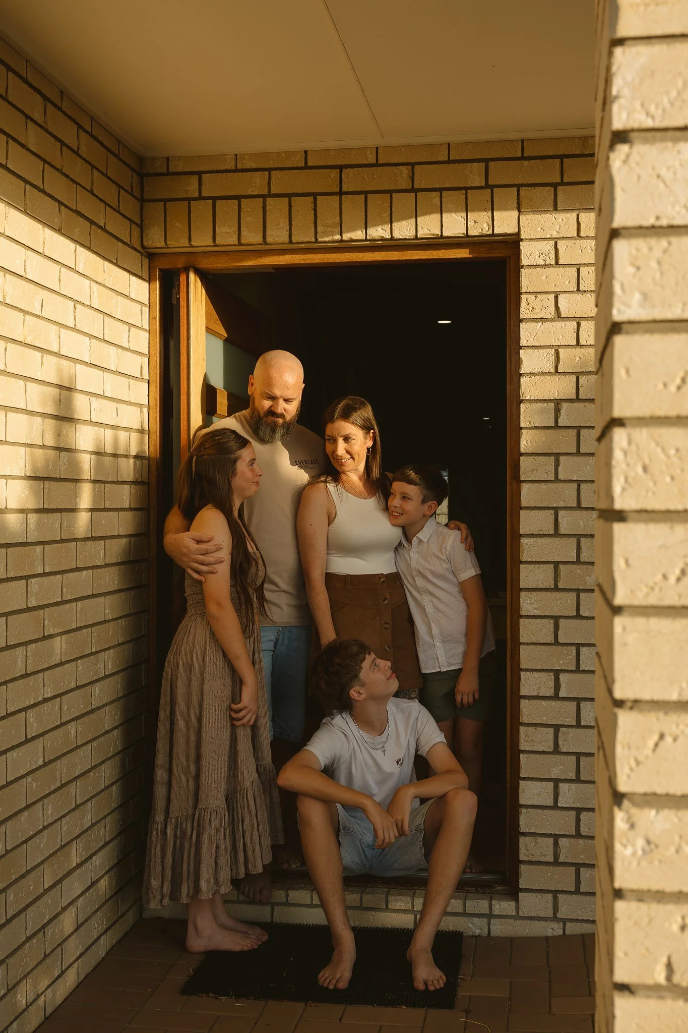 A group of six people, two adults and four children, are gathered together in a small space with brick walls. They are posing for a photo, with some standing and some sitting or crouching, all smiling and looking at each other or the camera.