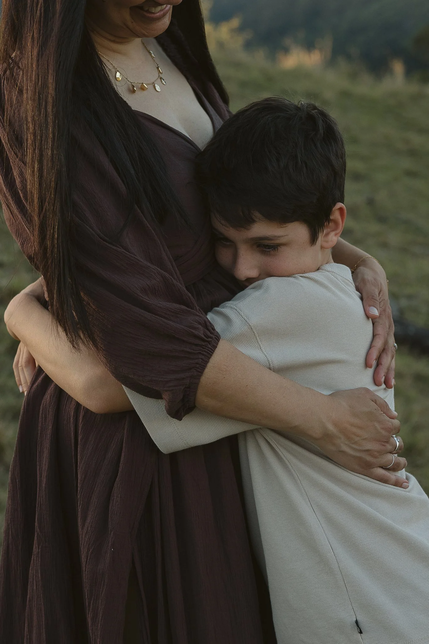 A woman with long dark hair and a necklace is hugging a young boy with short dark hair. They are outdoors in a grassy area at sunset.