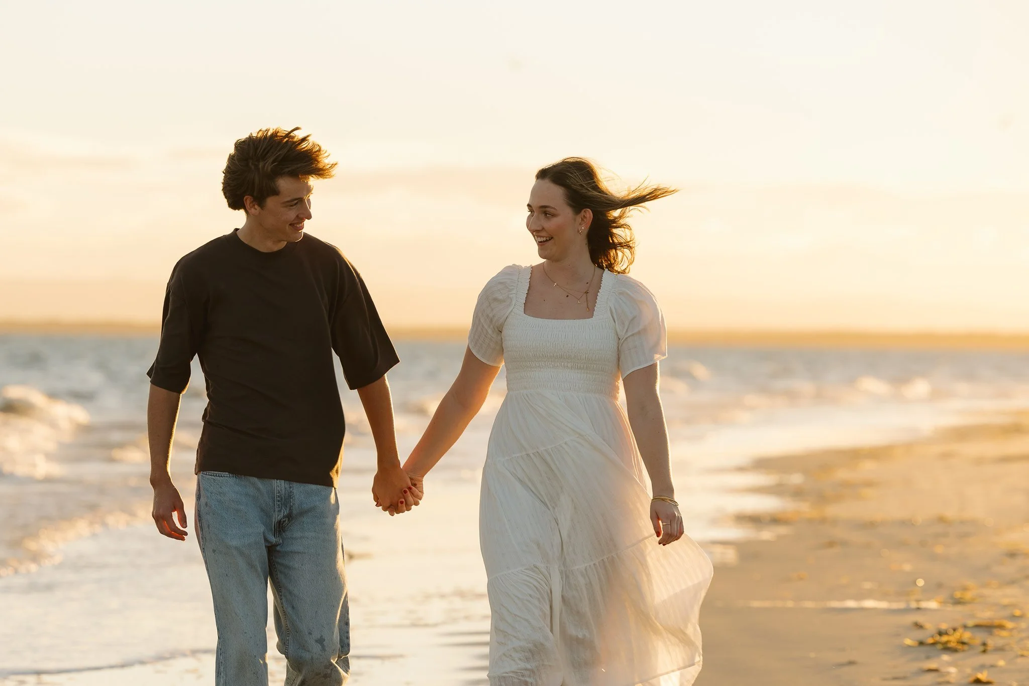 A couple walking hand in hand along the beach at sunset, smiling and enjoying each other's company.