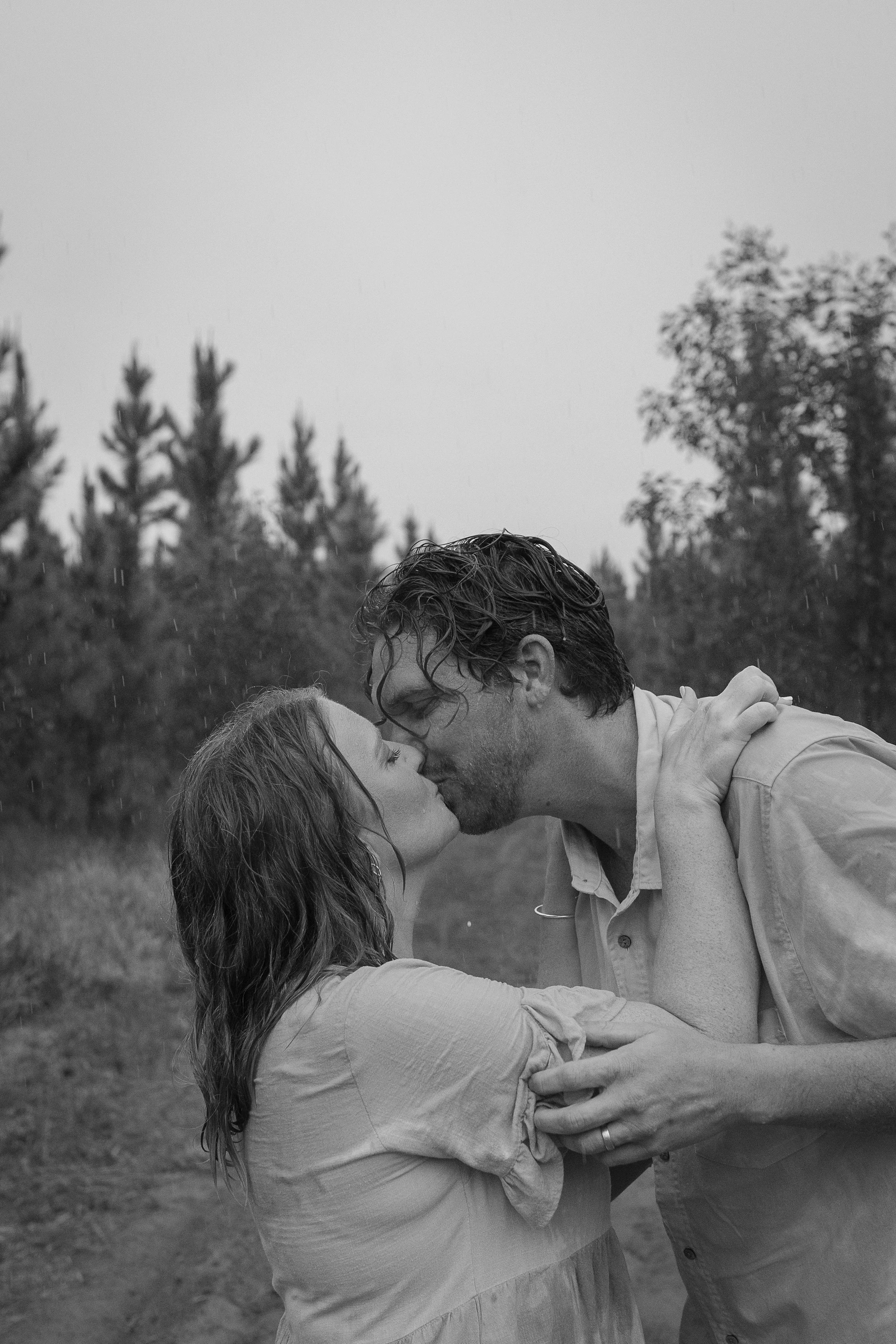 A couple is sharing an intimate moment outdoors on a rainy day, with the man gently holding the woman as they lean into each other, about to kiss, with trees in the background.
