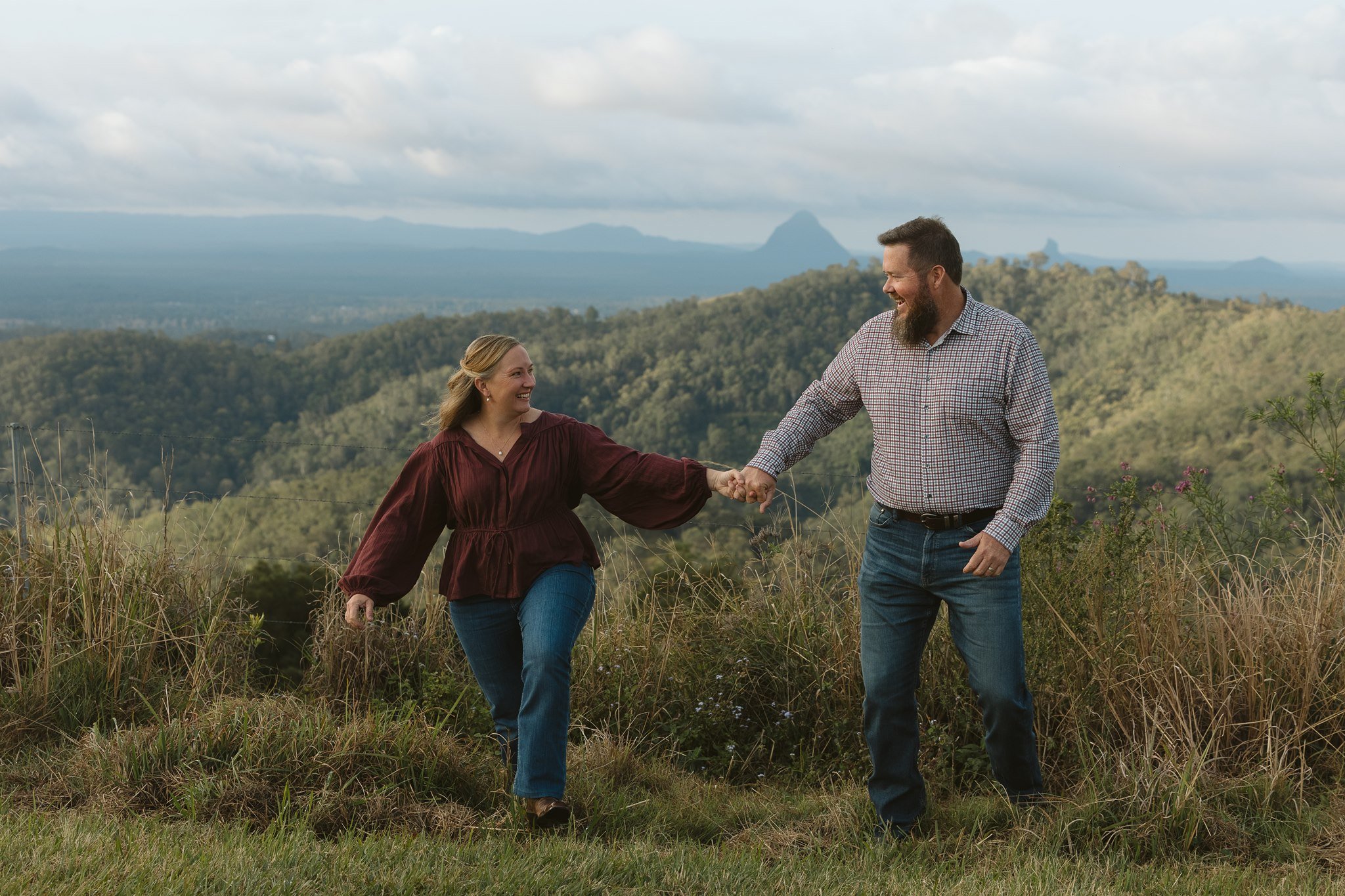 A man and a woman holding hands and smiling at each other outdoors with a scenic mountainous landscape in the background.