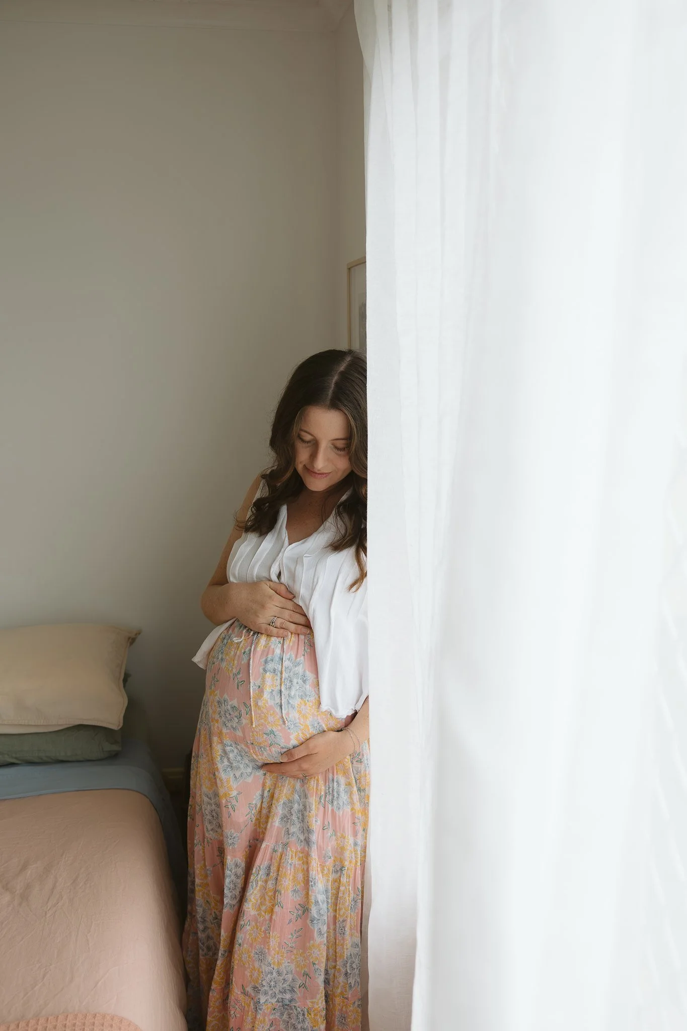 A pregnant woman with long brown hair holding her belly, standing in a room near a window with white sheer curtains. The woman is wearing a white top and a pink floral maxi skirt, and she is smiling softly looking down at her belly.