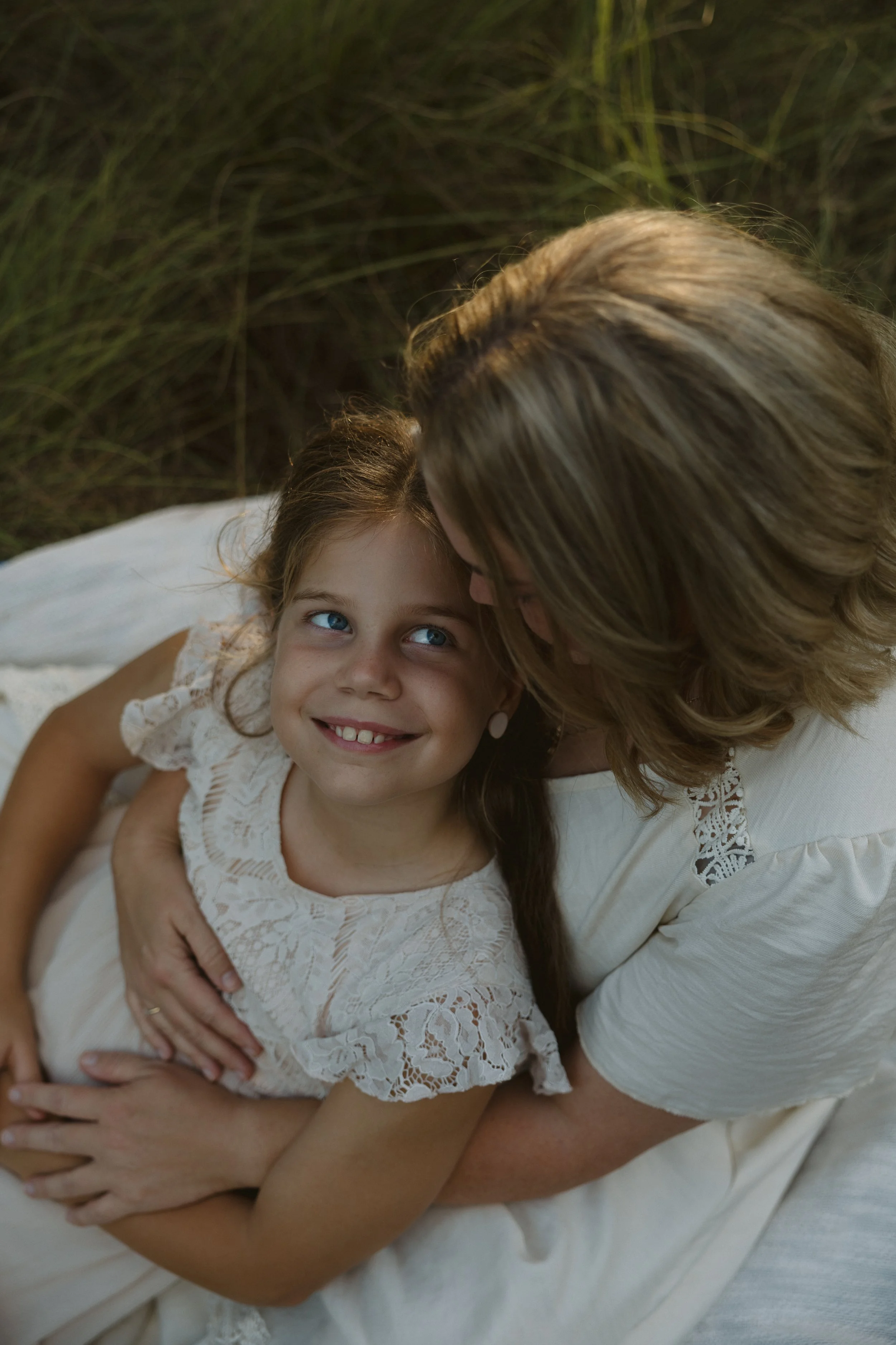 A woman with light brown hair embracing a young girl with brown hair and blue eyes while lying on a white bed outdoors in a grassy area.