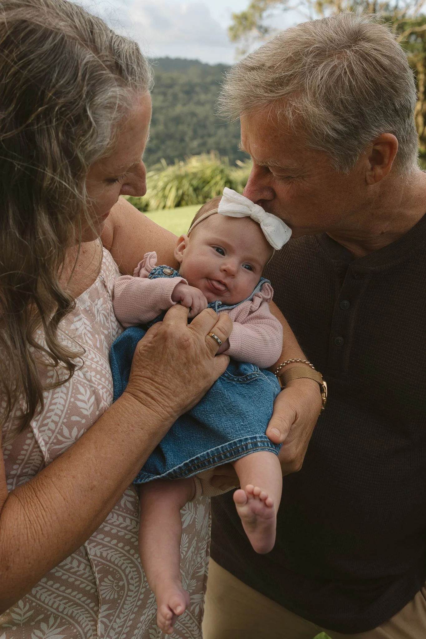 An elderly woman and man kiss a baby girl on the head outdoors, with mountains and greenery in the background.