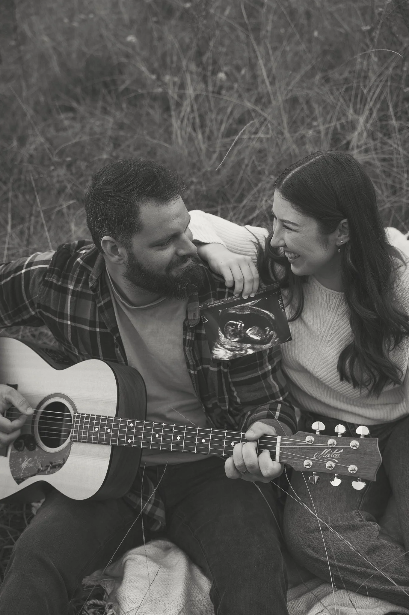 A man and woman sitting on grass outdoors, smiling at each other. The man is playing an acoustic guitar, and the woman is looking at him affectionately. The woman is holding an ultrasound image, indicating pregnancy. Both appear relaxed and happy.