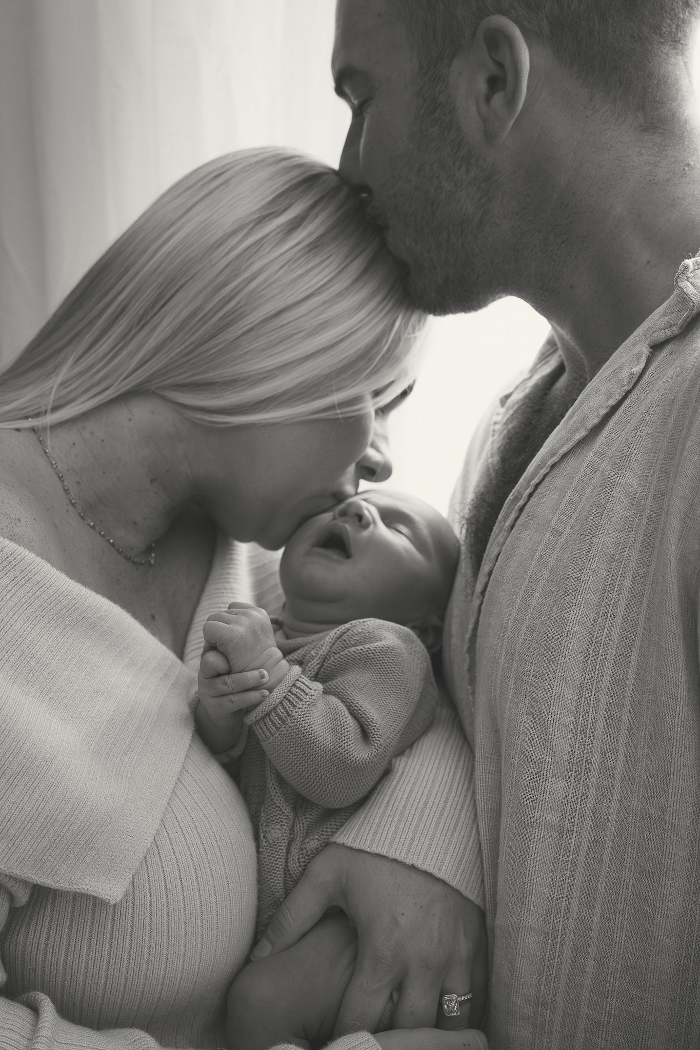 Black and white photo of a family of three: a man, a woman, and a baby. The woman is kissing the man on the forehead, and the baby is sleeping peacefully in the woman's arms, with the man's hand gently supporting the baby's head.
