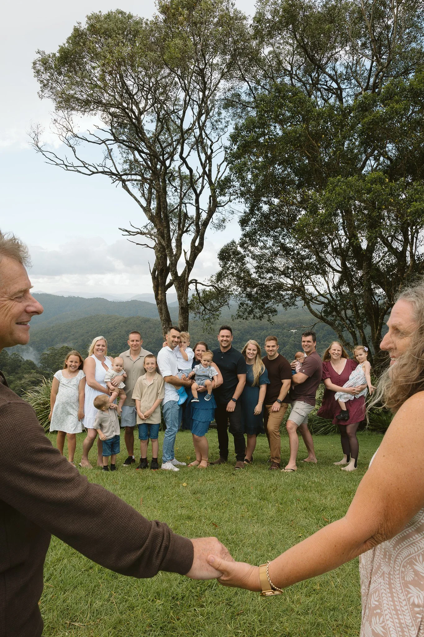 A group of people outdoors, holding hands and smiling, with mountains and trees in the background.