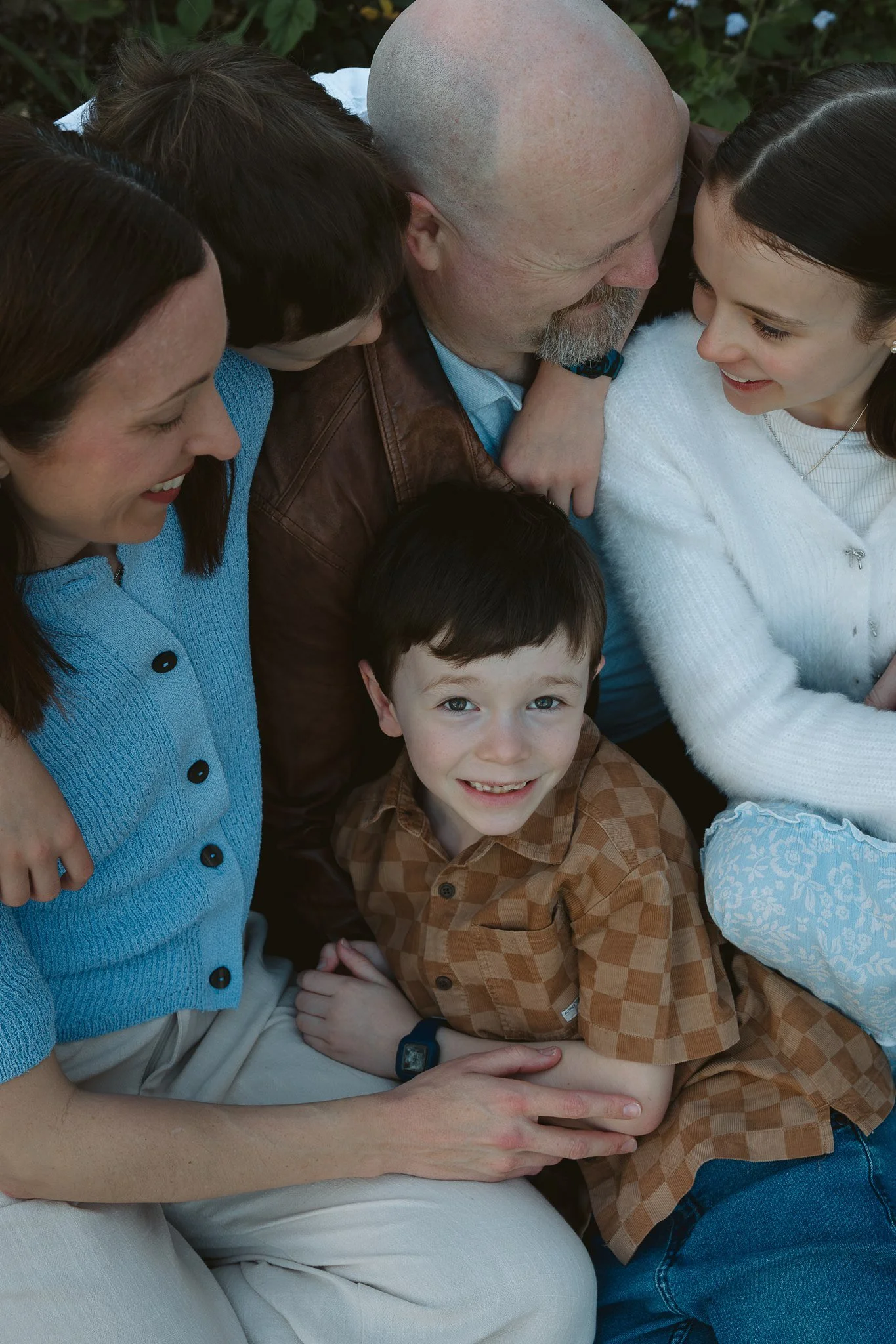 Group of six people, including three children and three adults, sitting closely together outdoors, smiling, with greenery in the background.