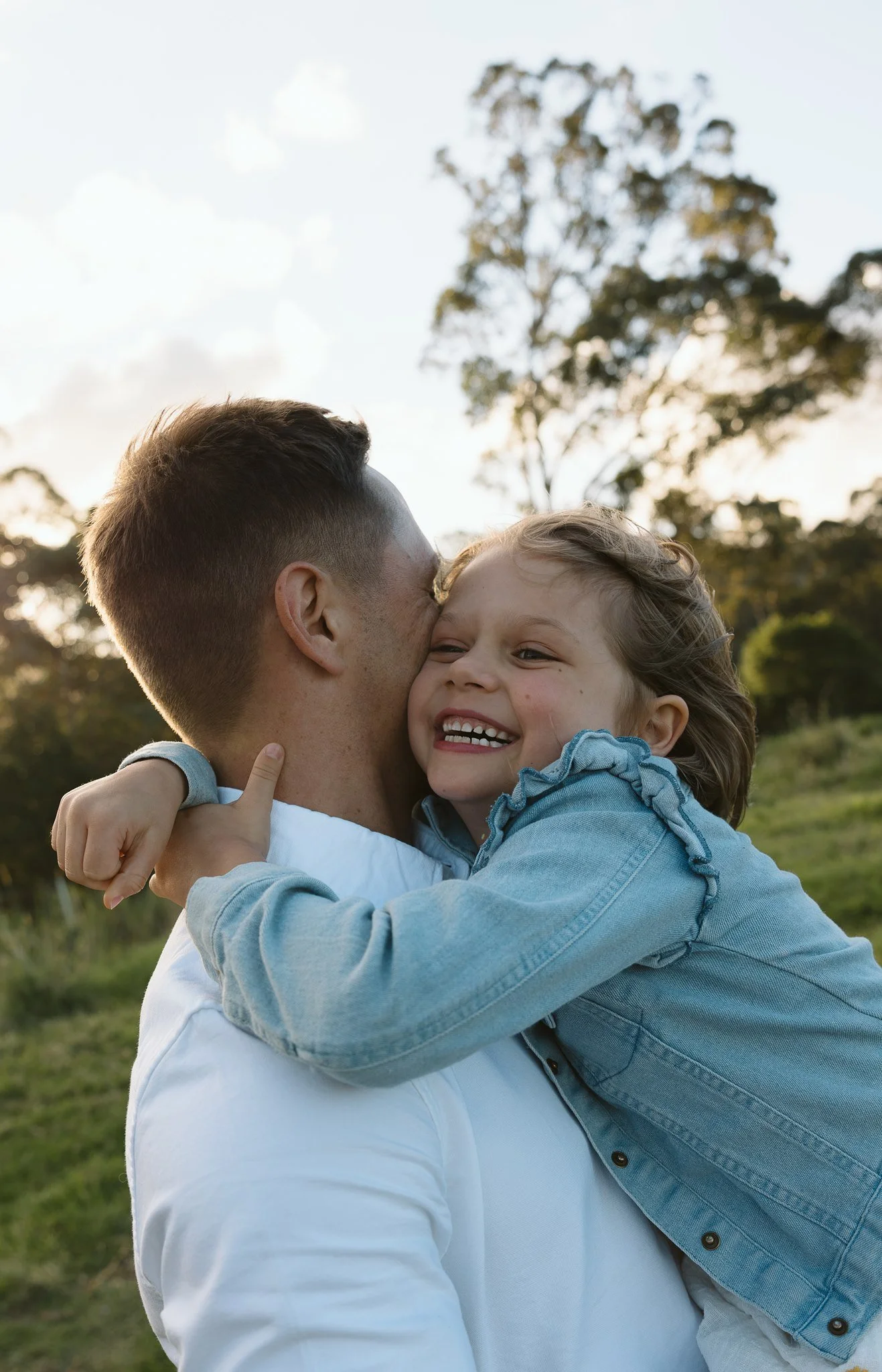 A man and a young girl are outdoors during daytime, sharing a joyful hug and smile. The girl is hugging the man tightly, and both appear very happy and affectionate. The background features trees and a partly cloudy sky.