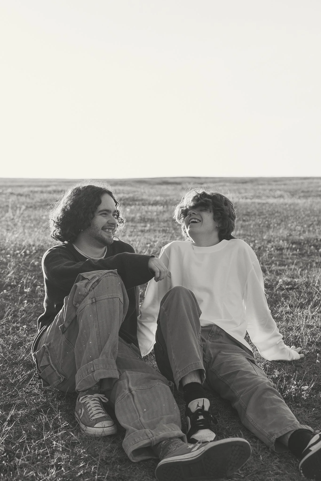 Two young men sitting on grass in a field, smiling and laughing, enjoying each other's company on a sunny day.