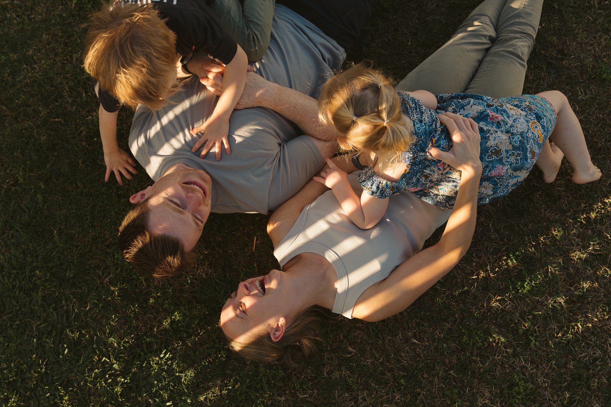 Family lying on grass, laughing, and hugging each other outdoors in sunlight.