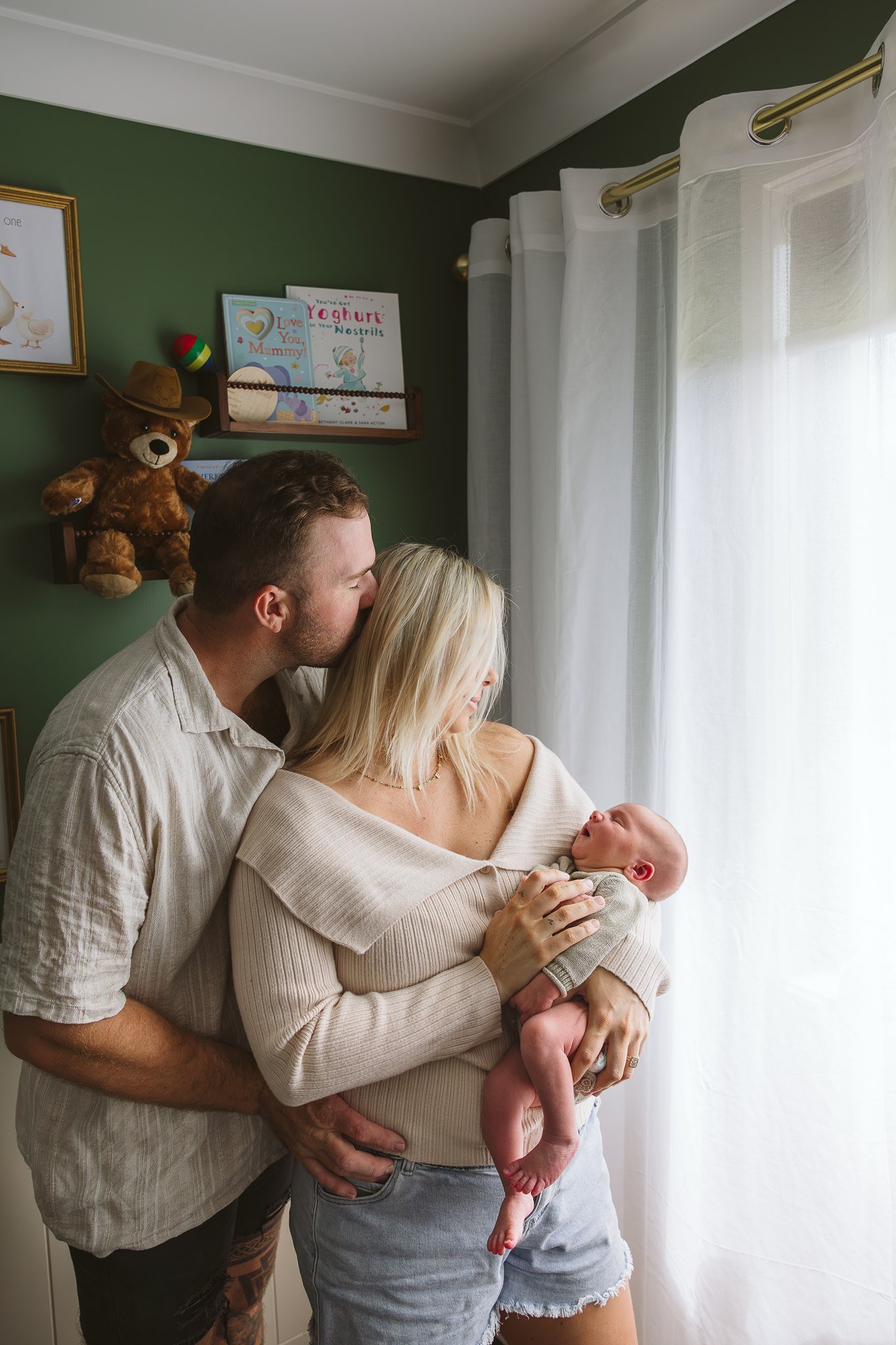 A couple holding their newborn baby during a moment of affection in a room with white curtains and framed pictures on a green wall, including a teddy bear with a hat and children's books.