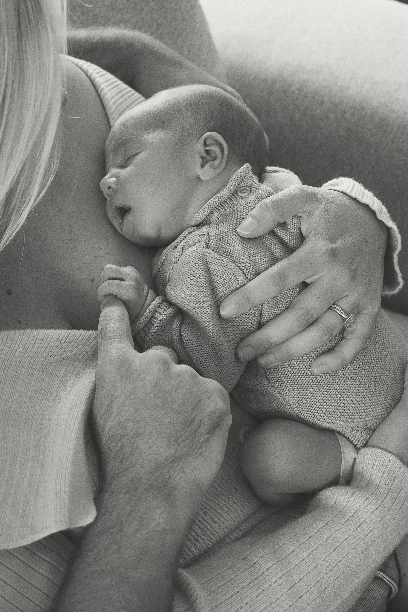 A baby sleeping on a mother's chest, with her hand gently holding the baby's head. The scene takes place indoors, with a soft surface and natural lighting.