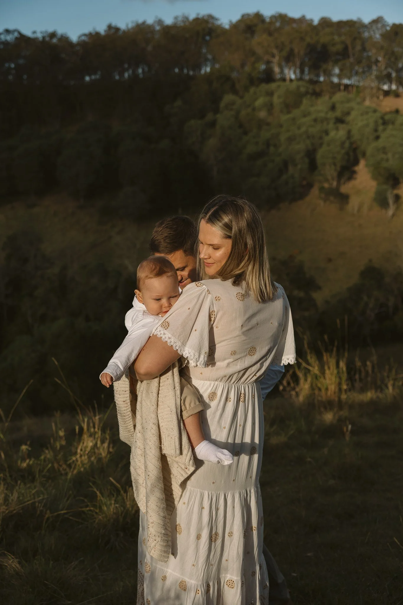 A woman holding a young child in a natural outdoor setting during golden hour. The scene features greenery, trees on a hill, and warm sunlight illuminating the subjects.