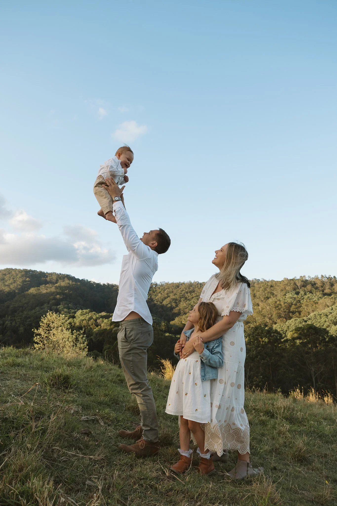 A family of five outdoors in a grassy field with trees and hills in the background. One man lifts a young boy into the air, and two women and a girl watch and smile. The sky is blue with some clouds.