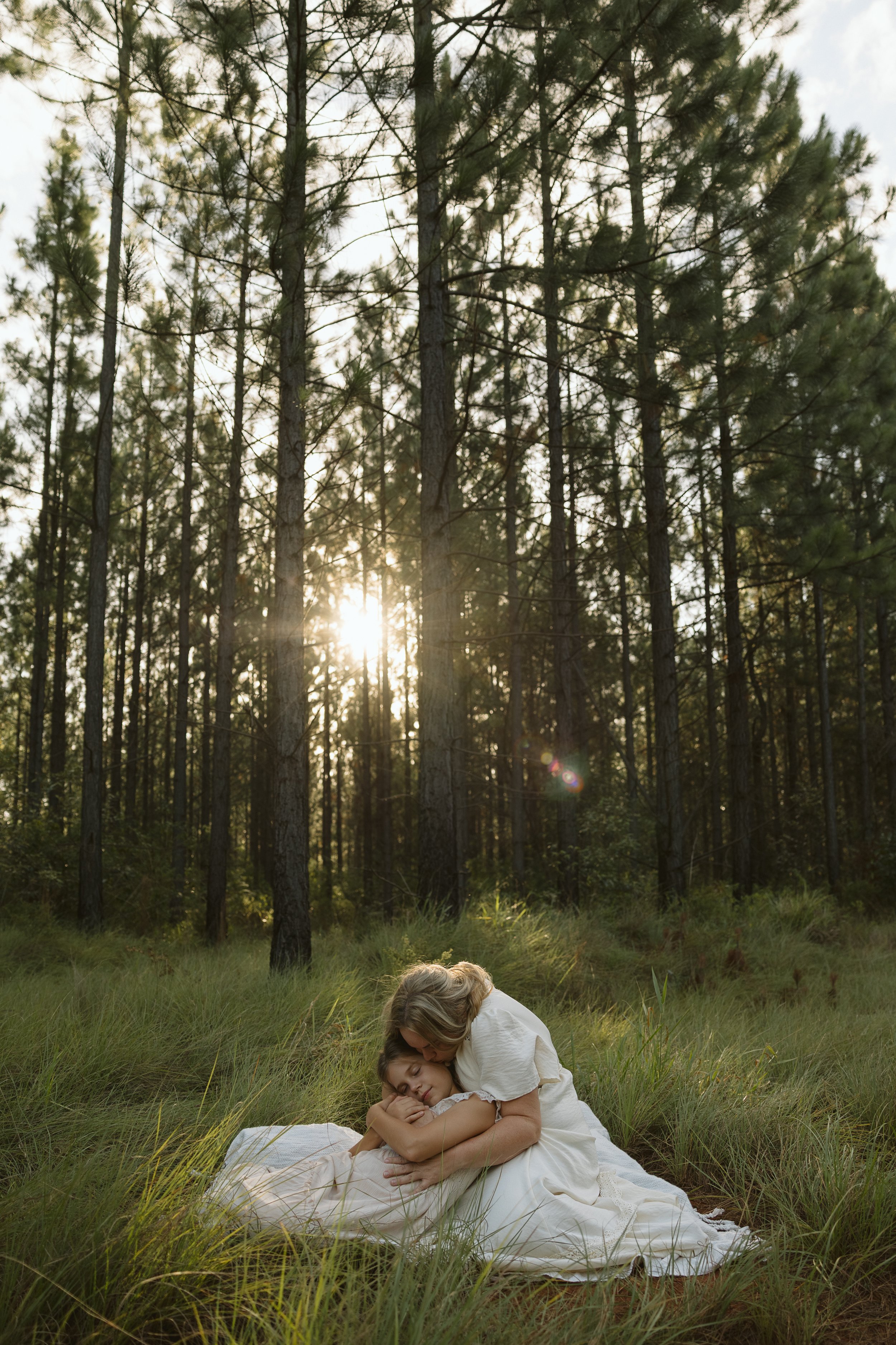 A woman and a girl embrace and lie on the grass in a forest clearing at sunset, with tall pine trees and sunlight filtering through the branches.