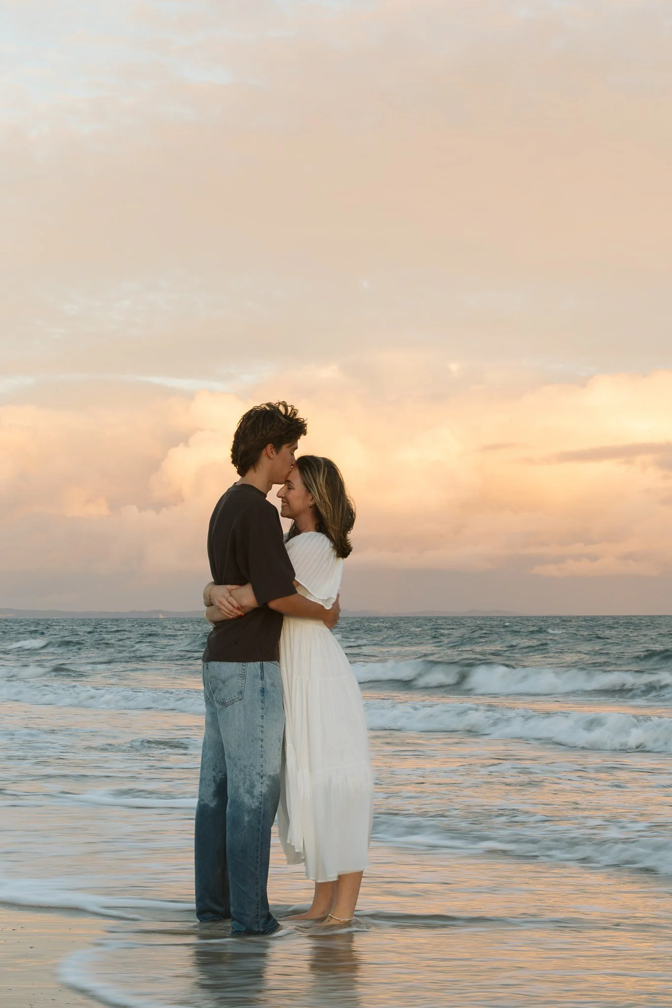 A young couple stands on the beach at sunset, embracing each other with the ocean in the background. The man is wearing a dark shirt and jeans, while the woman is in a white dress, both barefoot.