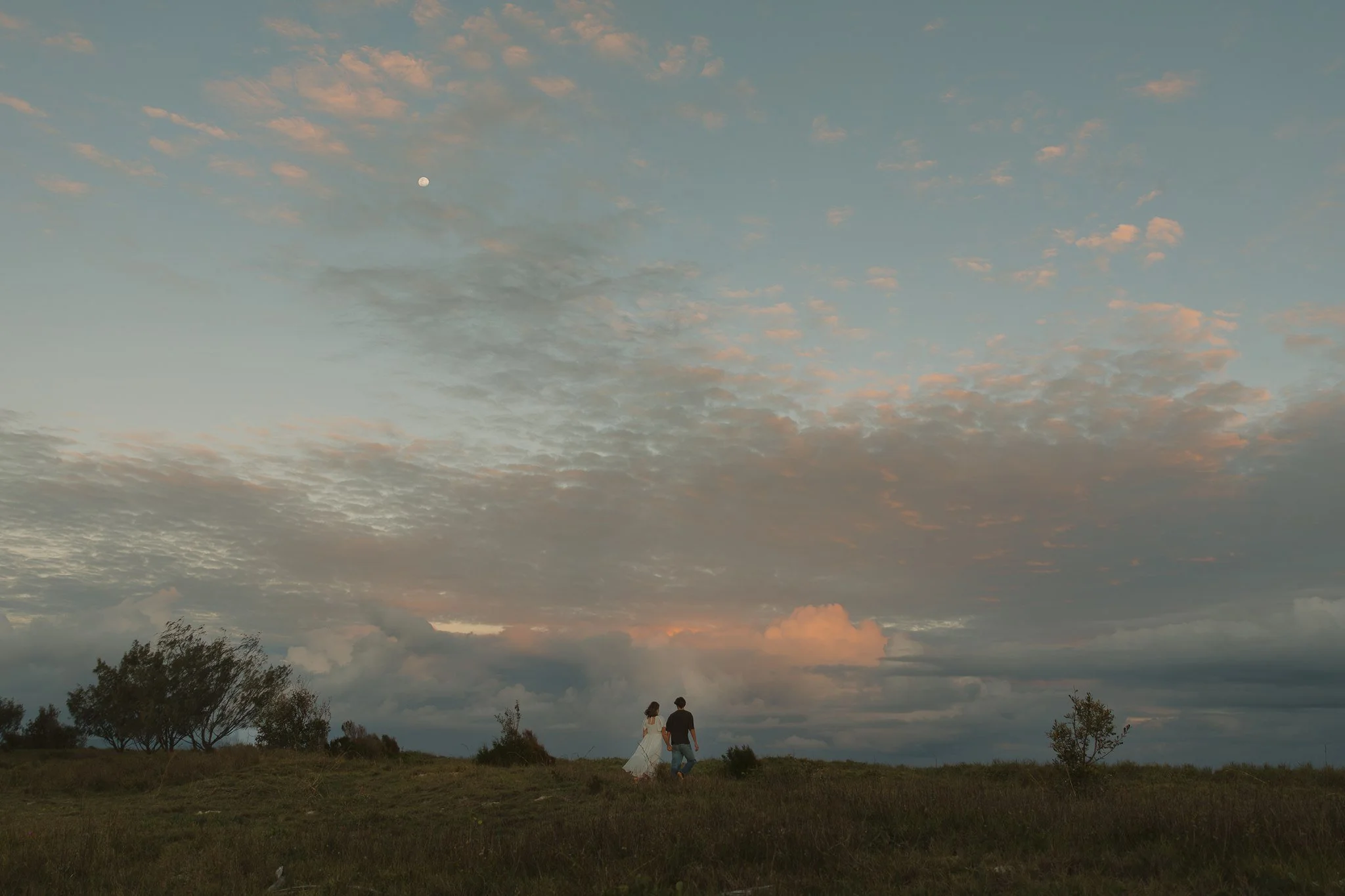 A couple walks hand in hand across a grassy field during sunset, with a cloudy sky and the moon visible overhead.