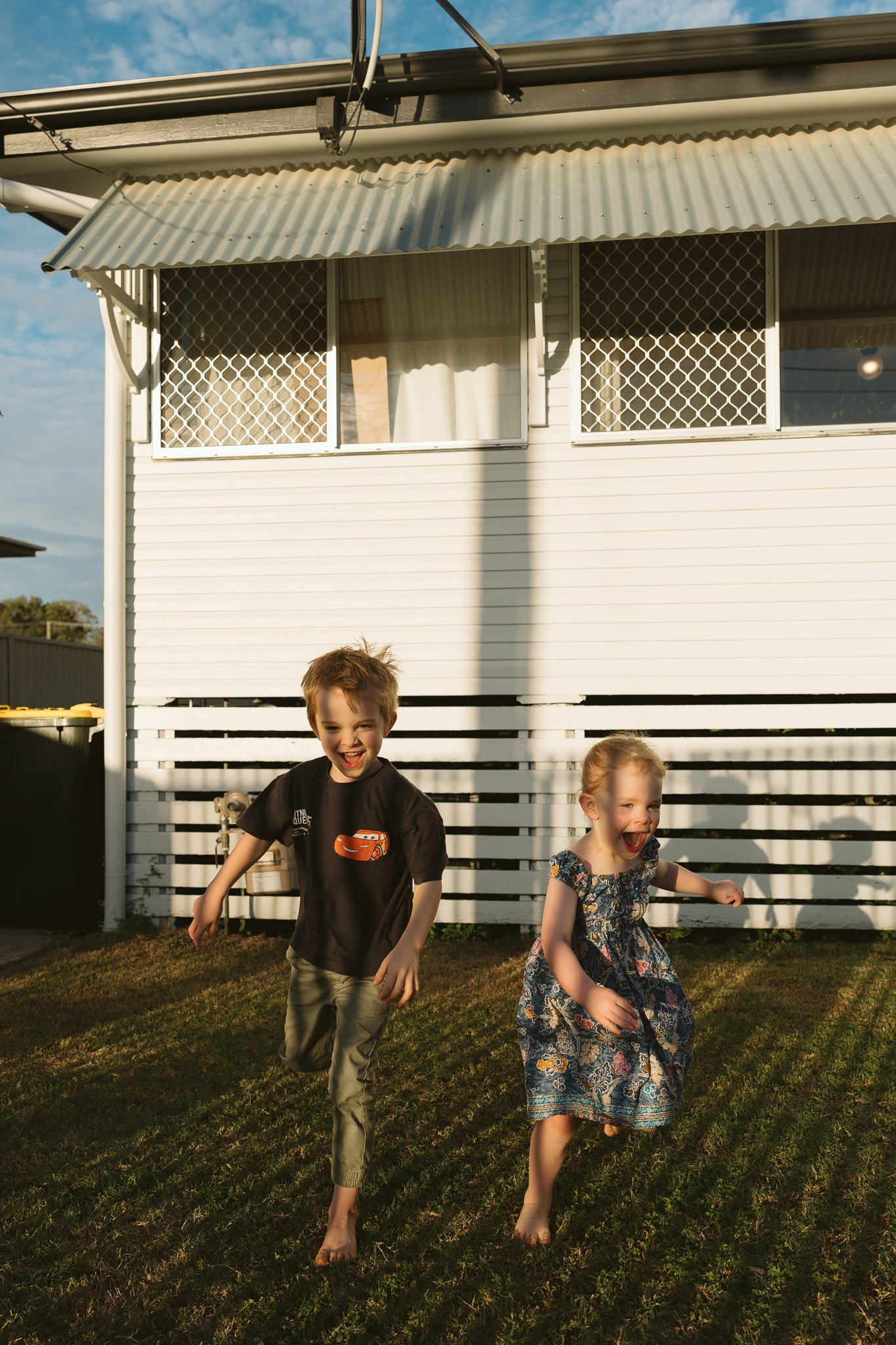 Two children, a boy and a girl, running barefoot on a lawn in front of a white house with a porch and a corrugated metal roof, during sunset.