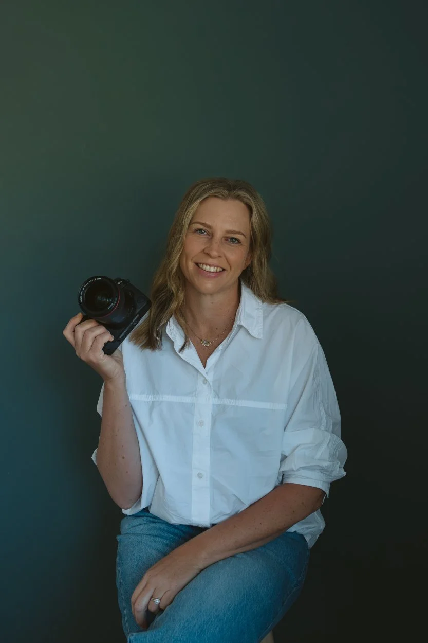 A woman with blonde hair, smiling, wearing a white button-up shirt and blue jeans, holding a camera, sitting against a dark background.