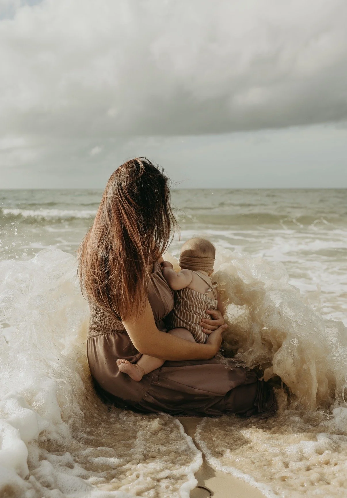 A woman holding a baby sitting on the beach near the ocean waves on a cloudy day.