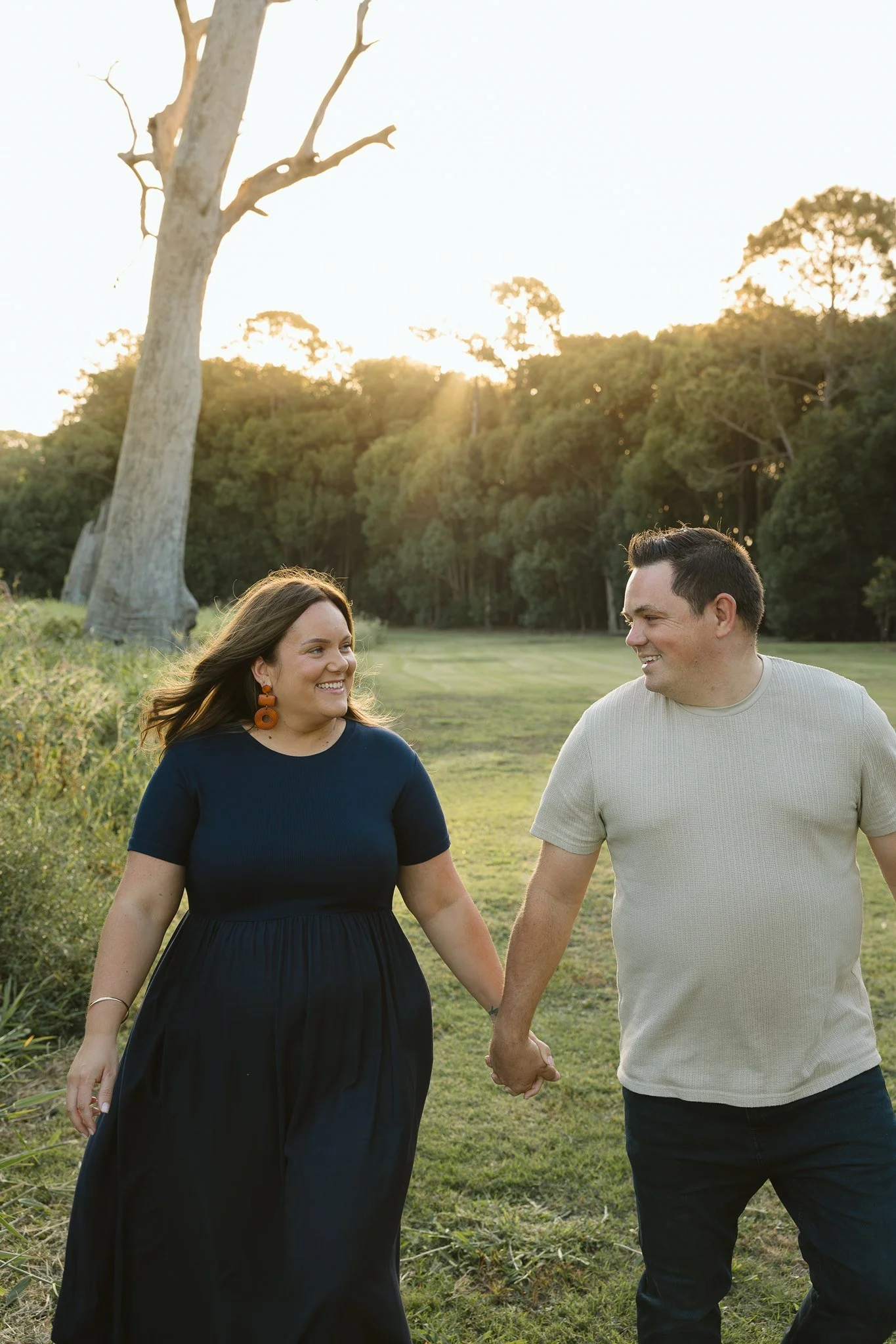 A smiling woman with brown hair, orange earrings, and a navy blue dress holding hands with a smiling man with short dark hair, wearing a light gray t-shirt, while walking outdoors in a grassy area with trees and a bright sky at sunset.
