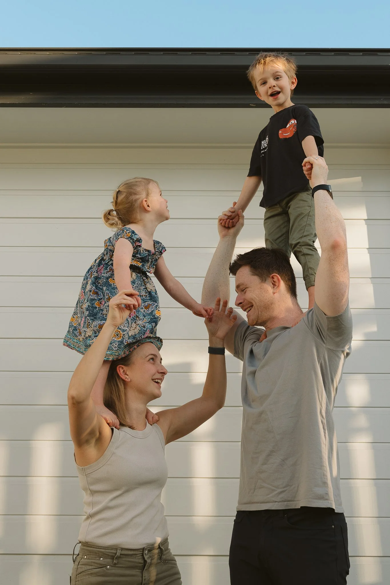 A family of four outside, with a man holding a young boy on his shoulders, a woman holding a girl on her shoulders, against a white wall and clear sky.