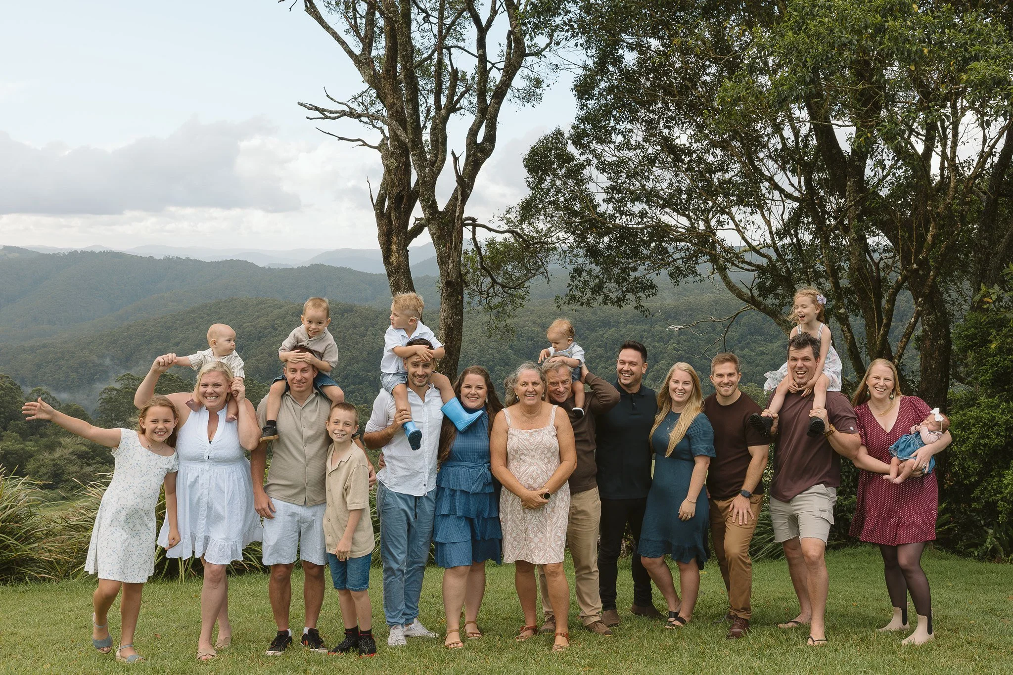 A large group of people, including children and adults, standing outdoors in front of a scenic mountainous landscape with trees, during daytime.