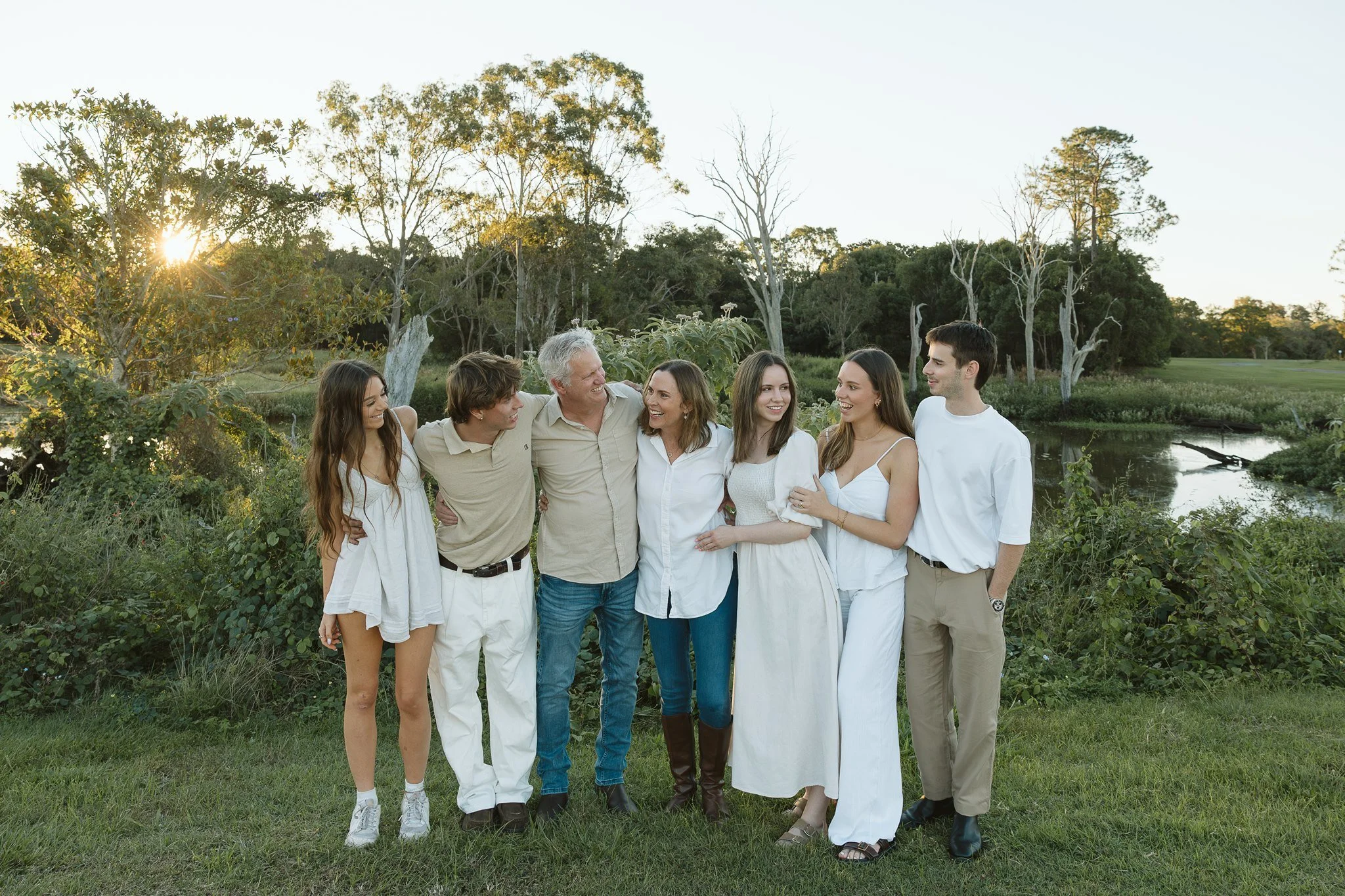 A group of seven people, including young adults and an older man, standing outdoors near a river, smiling and enjoying each other's company during sunset.