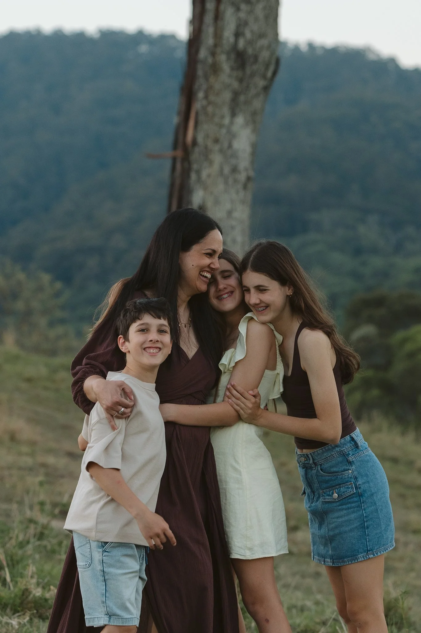 A woman with three children smiling and embracing outdoors with green hills and a large tree in the background.