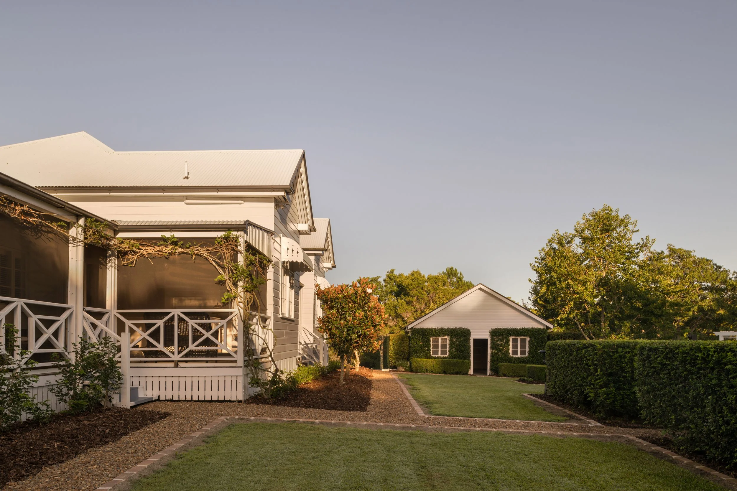 View of a backyard with a well-maintained lawn, a paved walkway, and various trees and bushes. There are residential buildings with white siding, balconies, and screened porches. The sky is clear with no clouds.