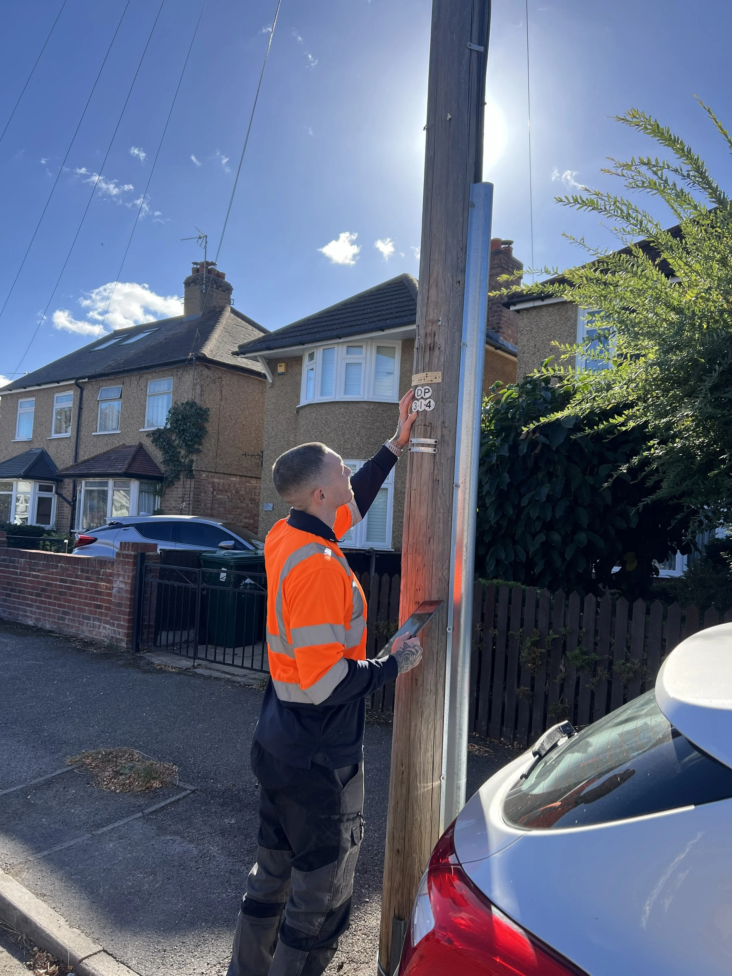 A man wearing an orange and gray jacket working on a wooden utility pole during sunny weather. The man has a tattoo on his left wrist and is holding a tablet device. Residential houses and cars are visible in the background.