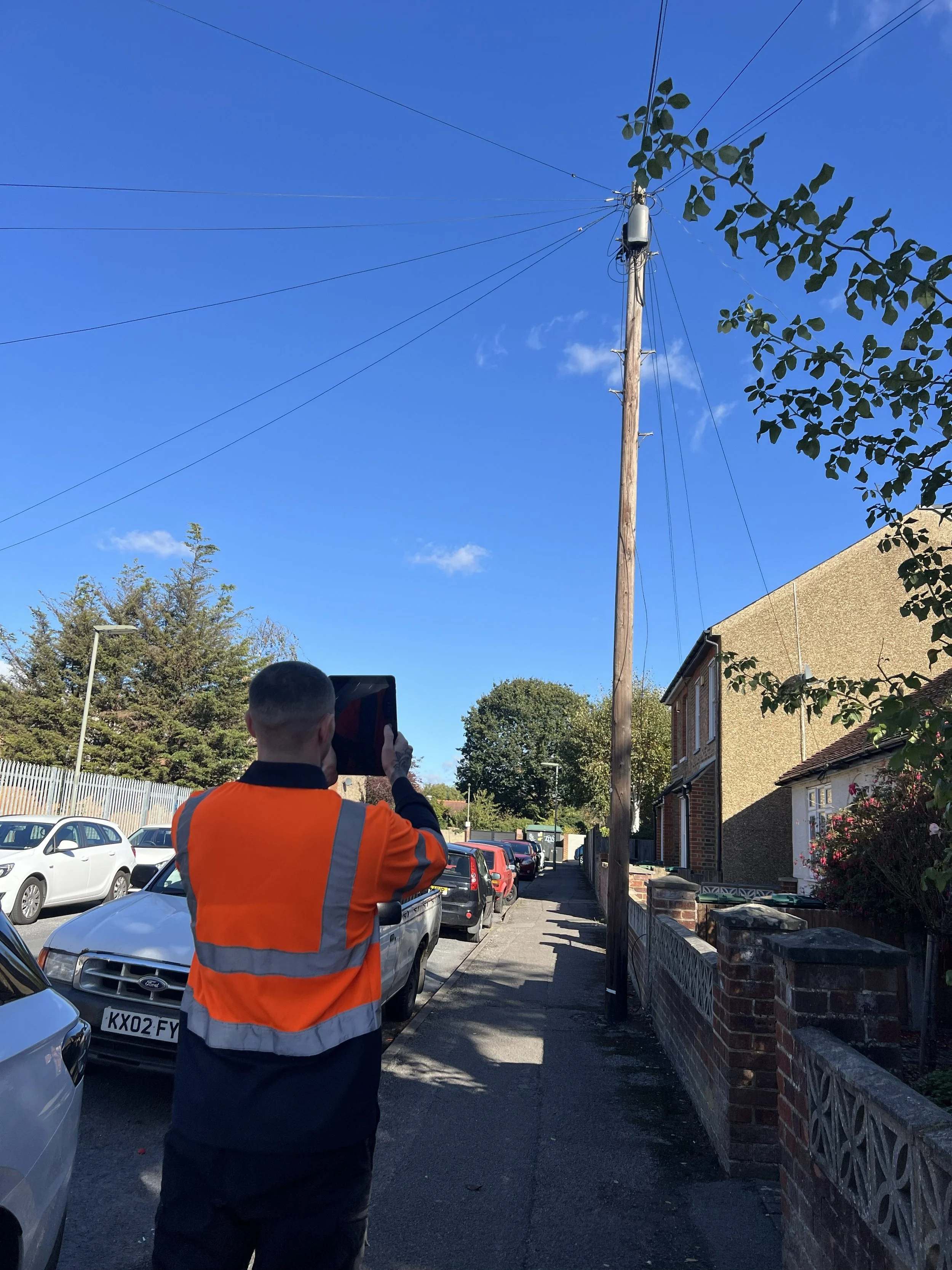 A person in an orange safety vest is taking a photo of a wooden utility pole with electrical wires on a residential street under a blue sky with some clouds.
