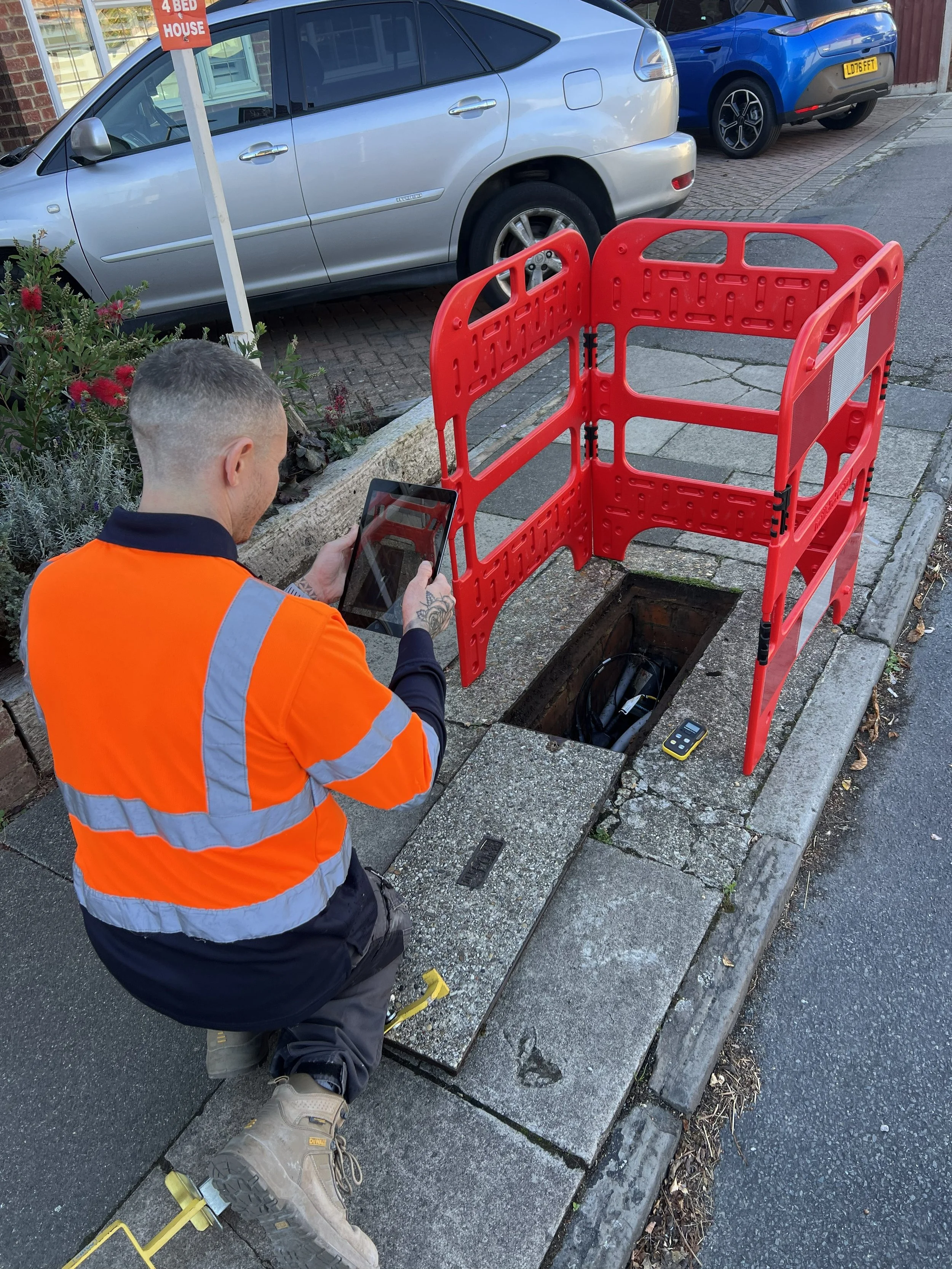 A worker wearing a bright orange safety vest and khaki work boots kneels next to an open manhole on the sidewalk. A red safety barrier surrounds the opening, and the worker is using a tablet or device. Parked cars line the street behind him, including a silver car and a blue car, with a residential brick building visible in the background.