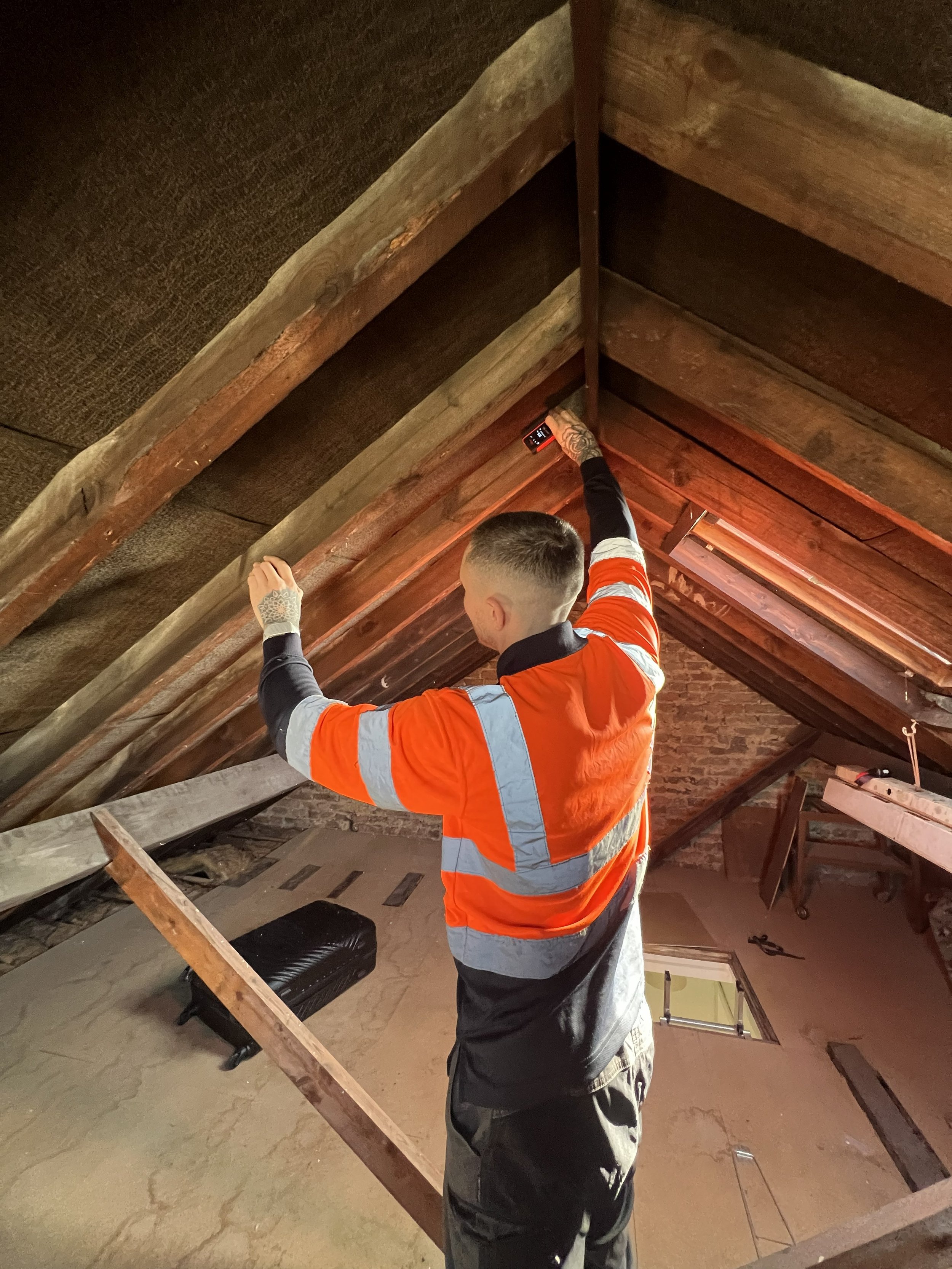 A person wearing an orange safety jacket and dark pants standing under a sloped wooden ceiling, holding a small electronic device, possibly a smartphone, with one arm extended upward.