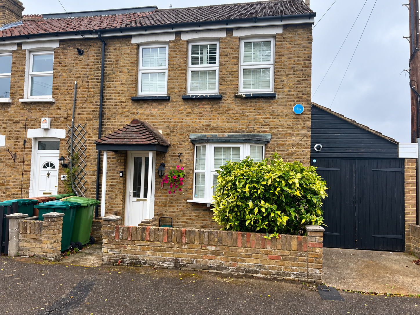 A brick house with white-framed windows, a small bay window, and a front door with a tiled porch. There is a green shrub in front, hanging flower baskets, and a black garage door to the right. Recycling bins are on the sidewalk.
