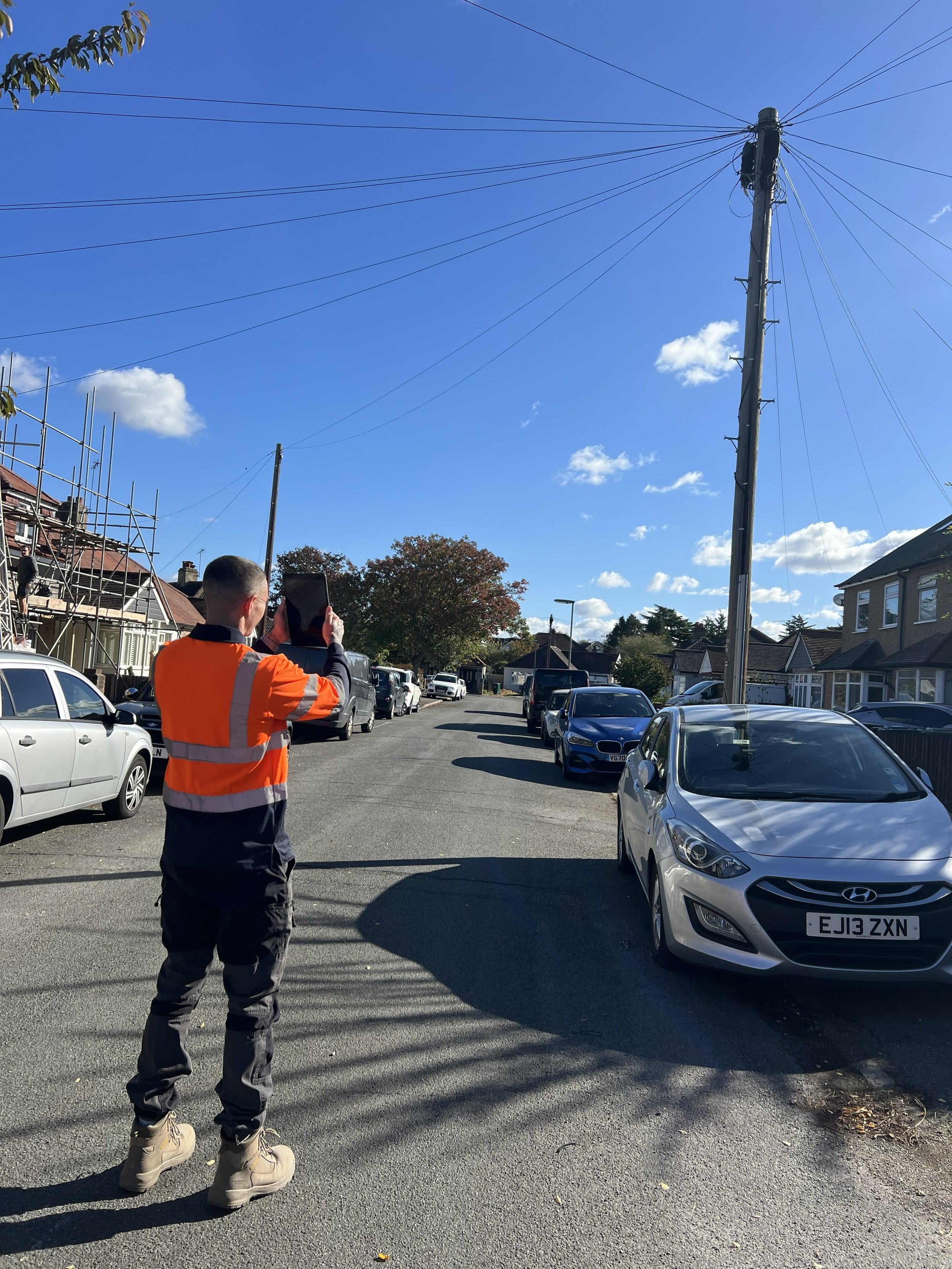 A man in a high-visibility orange jacket and black pants stands on a city street taking a photo with his phone. Parked cars line both sides of the street, and houses with tiled roofs are visible in the background. A tall utility pole with power lines is prominent in the scene, set against a bright blue sky with some scattered clouds.
