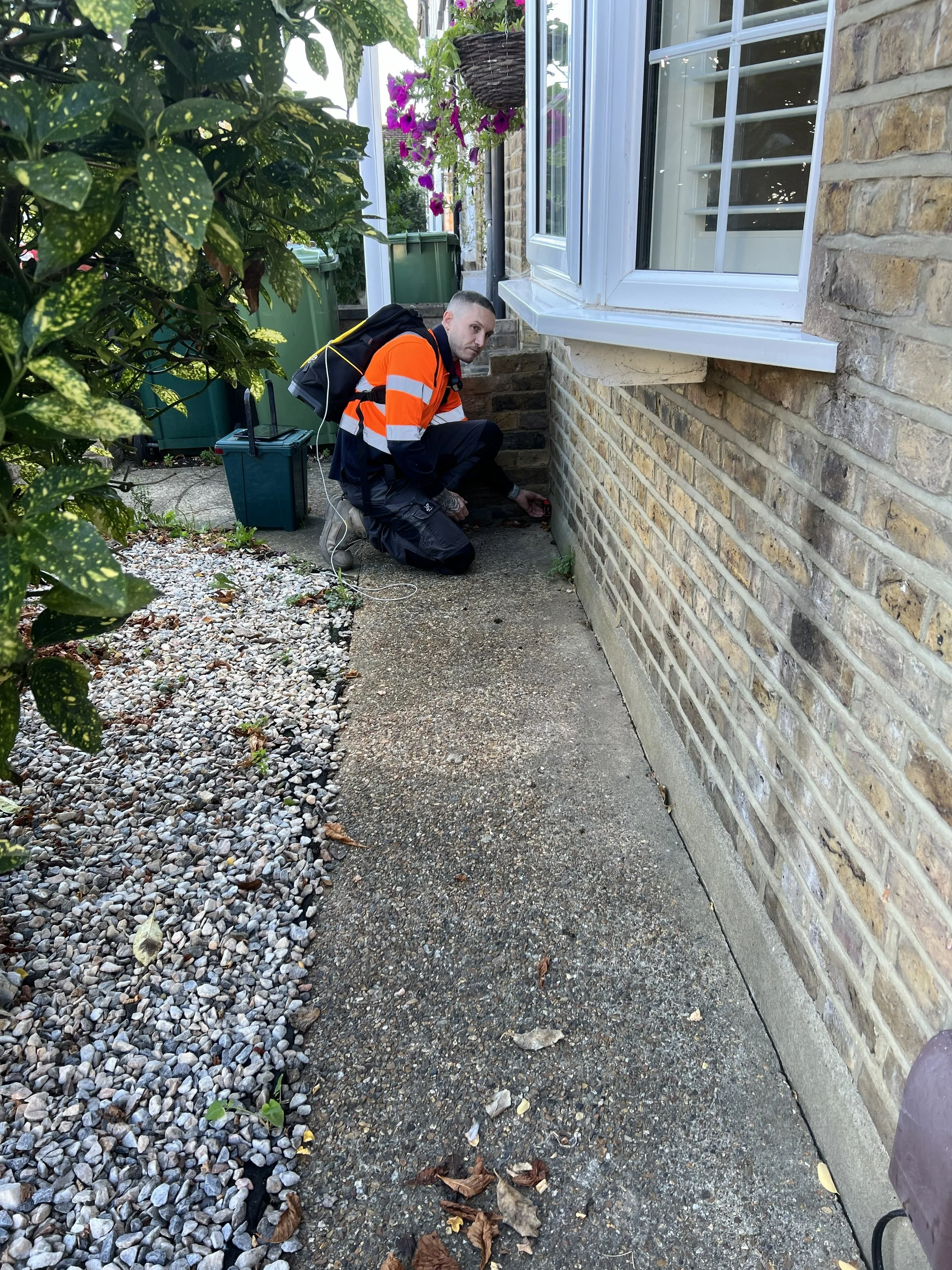 A man kneeling outside next to a brick house, working on the foundation, with a backpack, electronic device, and tools nearby. There are plants, flowers, and bushes around.