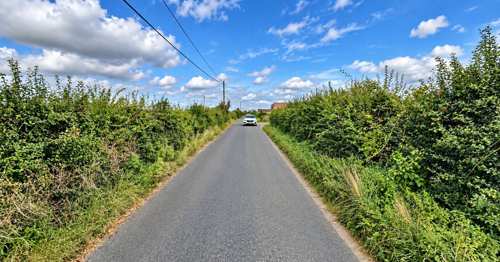 A narrow country road with a white car driving towards the camera, bordered by dense green bushes and trees on both sides, under a partly cloudy blue sky with scattered white clouds.